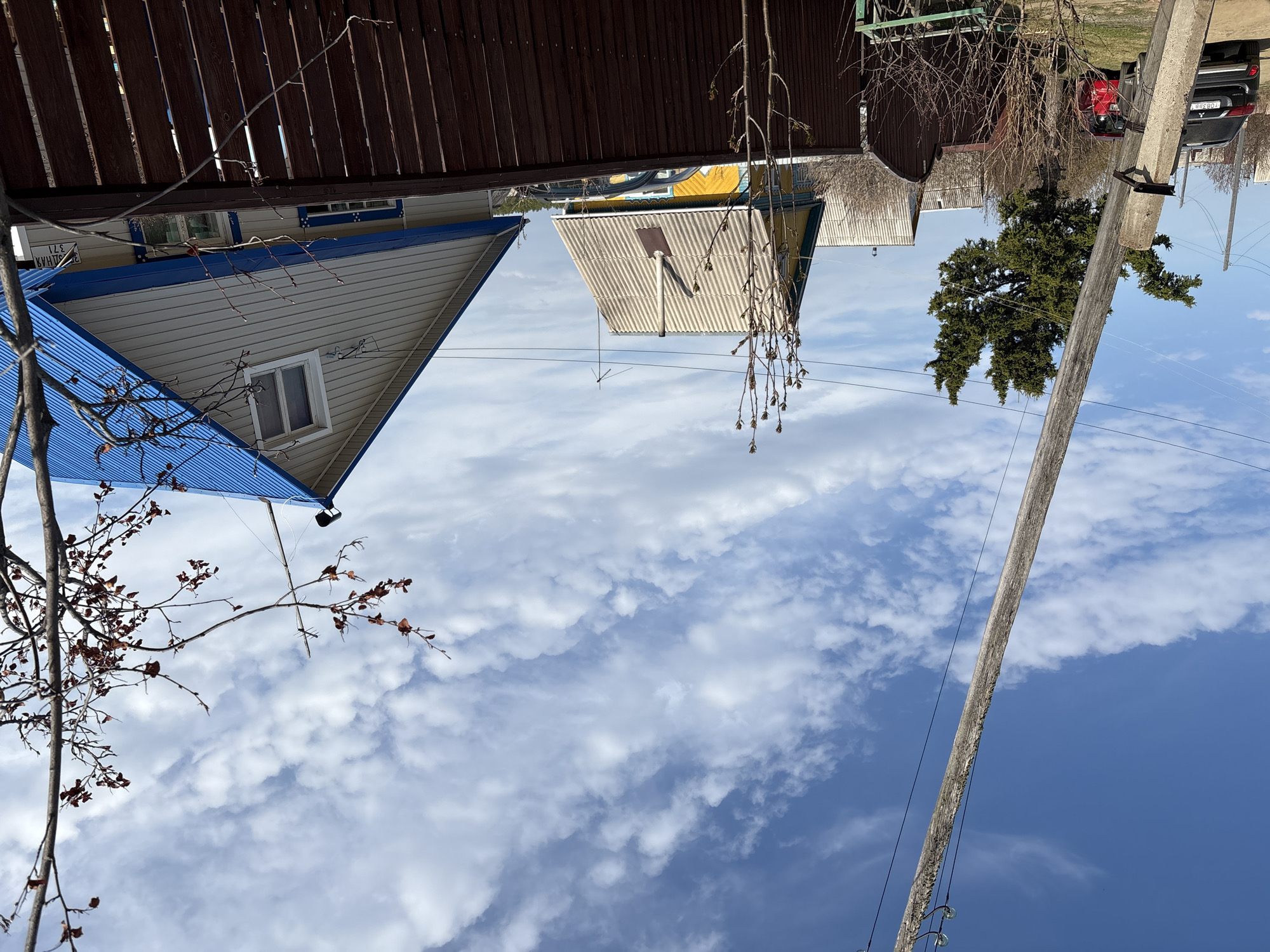 The image captures a rural or suburban residential scene under a clear sky with scattered white clouds. The perspective is slightly upwards, looking over fences and houses towards the horizon.
In the foreground, on the left, bare tree branches with some dried, reddish leaves are visible. To the right of these branches, the gable end of a house with light-colored siding and a bright blue corrugated metal roof is prominent. A single window is centered on this gable. Above the window and partially obscured by a dark wooden fence, Cyrillic text reading "КВАРТИРЫ" (Apartments) and "371" is visible on the house's exterior. The fence, made of dark brown wooden planks, runs horizontally across the upper-left portion of the image, appearing to separate properties.
Further into the scene, another house with a lighter-colored corrugated metal roof (possibly white or light yellow) and greyish siding is seen. A small, dark, shovel-like object or antenna is mounted on its roof. More bare tree branches with small buds or dried leaves are in front of this house.
Towards the right side of the image, a large, dark green coniferous tree stands. A sturdy wooden utility pole, equipped with insulators and power lines, extends vertically upwards on the far right. Behind the tree and pole, parts of a red car and a black SUV are visible. The black SUV's license plate displays "T083BB" and region code "138".
The weather appears to be bright and clear daytime, with blue sky dominating the upper right portion and transitioning to a lighter blue with white, puffy clouds towards the left. There is no specific activity taking place; the scene depicts a static outdoor environment.