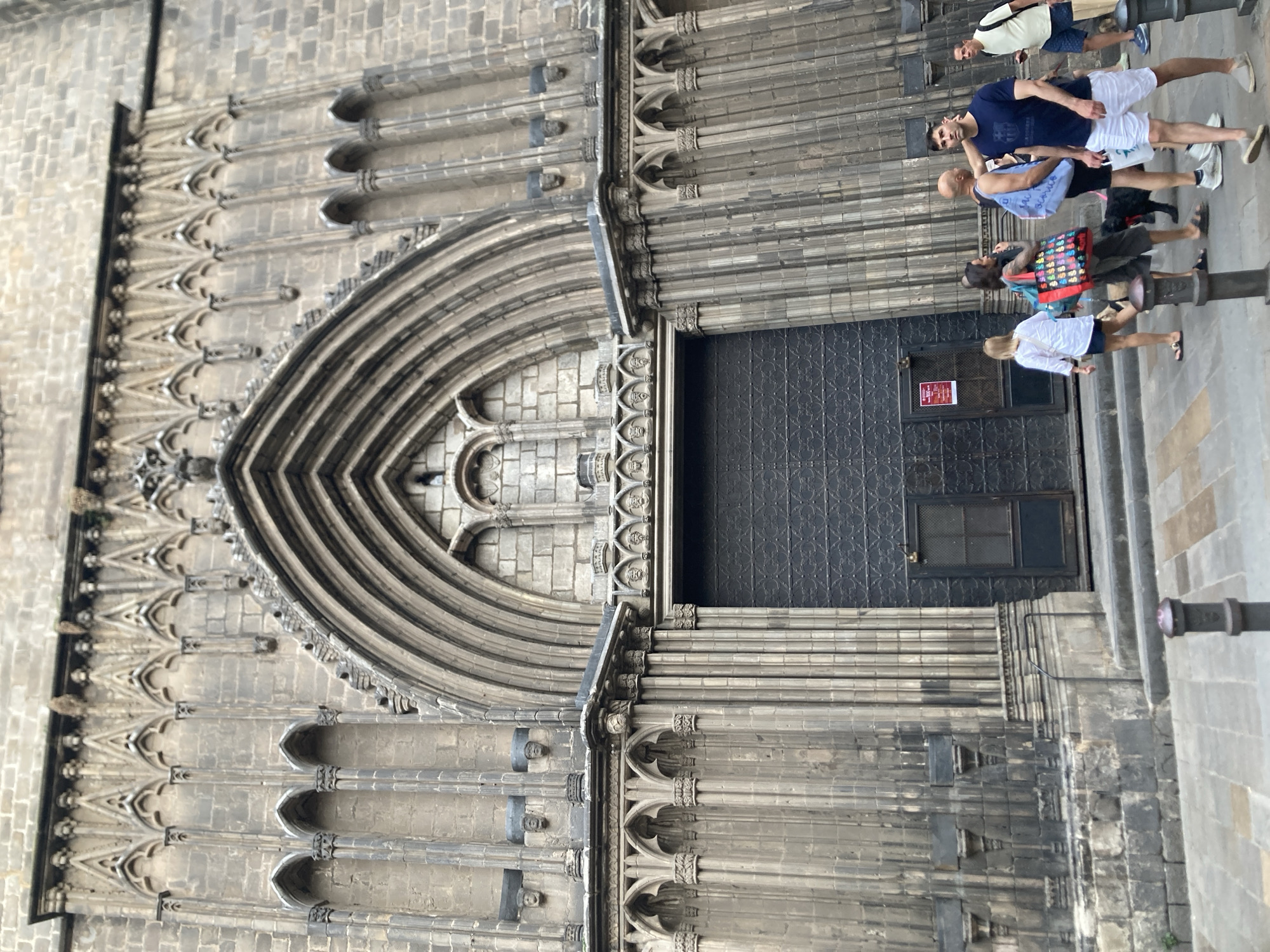 The image shows the imposing facade of the Barcelona Cathedral, located in the Gothic Quarter of Barcelona, Spain. The architecture is characterized by intricate stone carvings, pointed arches, and ornate decorative elements. A large, dark metal door is centered within a grand arched entrance. Several people are visible in front of the cathedral, some walking and others standing. They appear to be tourists or locals. The ground in front of the cathedral is paved with cobblestones. The lighting suggests it is daytime, with some shadows indicating the sun's position. The overall impression is one of historical grandeur and a bustling urban environment.