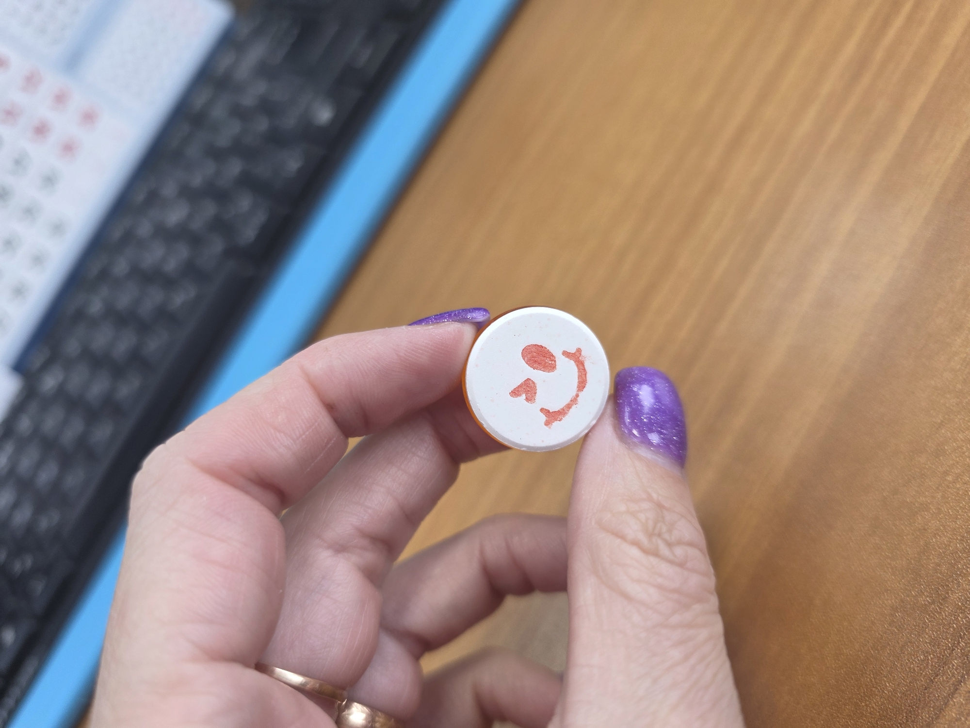 A hand holds a white, circular object with a red smiling face drawn on it. The hand has a gold ring on one finger and the fingernails are painted purple with glitter. The background is a wooden surface and a blue object is visible to the side. The image was taken in Chaikovsky, Russia.