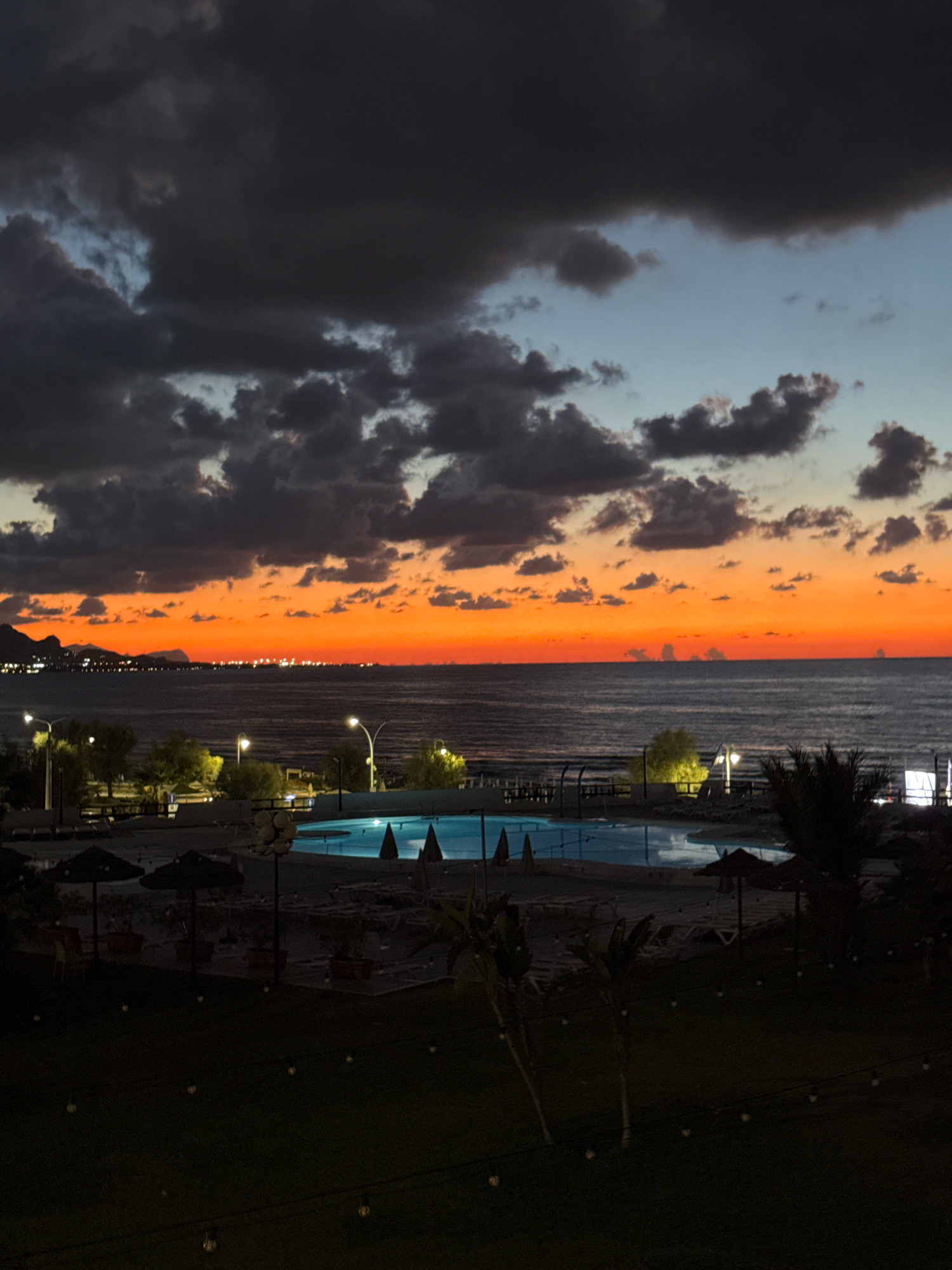 A twilight view captures a coastal resort area in Isola delle Femmine, Italy, with a dramatic sunset painting the sky. The horizon is dominated by a vivid gradient of orange and red, transitioning to dusky blues and grays as dark, heavy clouds gather overhead. The Tyrrhenian Sea stretches out, its surface dark and slightly rippled, reflecting the fading light.
In the foreground, a resort area is visible. A brightly lit swimming pool, with its blue water contrasting against the twilight, is a central feature. Around the pool, lounge chairs and umbrellas are arranged, some casting long shadows. Scattered vegetation, including palm trees and smaller plants, adds to the resort ambiance. The ground in the immediate foreground is covered in grass, with strings of small, illuminated fairy lights adding a festive touch. In the distance, along the coastline, the lights of a town or city are visible, hinting at the settlement of Isola delle Femmine. No people are explicitly visible in the frame, suggesting a peaceful, late-evening atmosphere. The overall impression is one of serene beauty as day gives way to night.