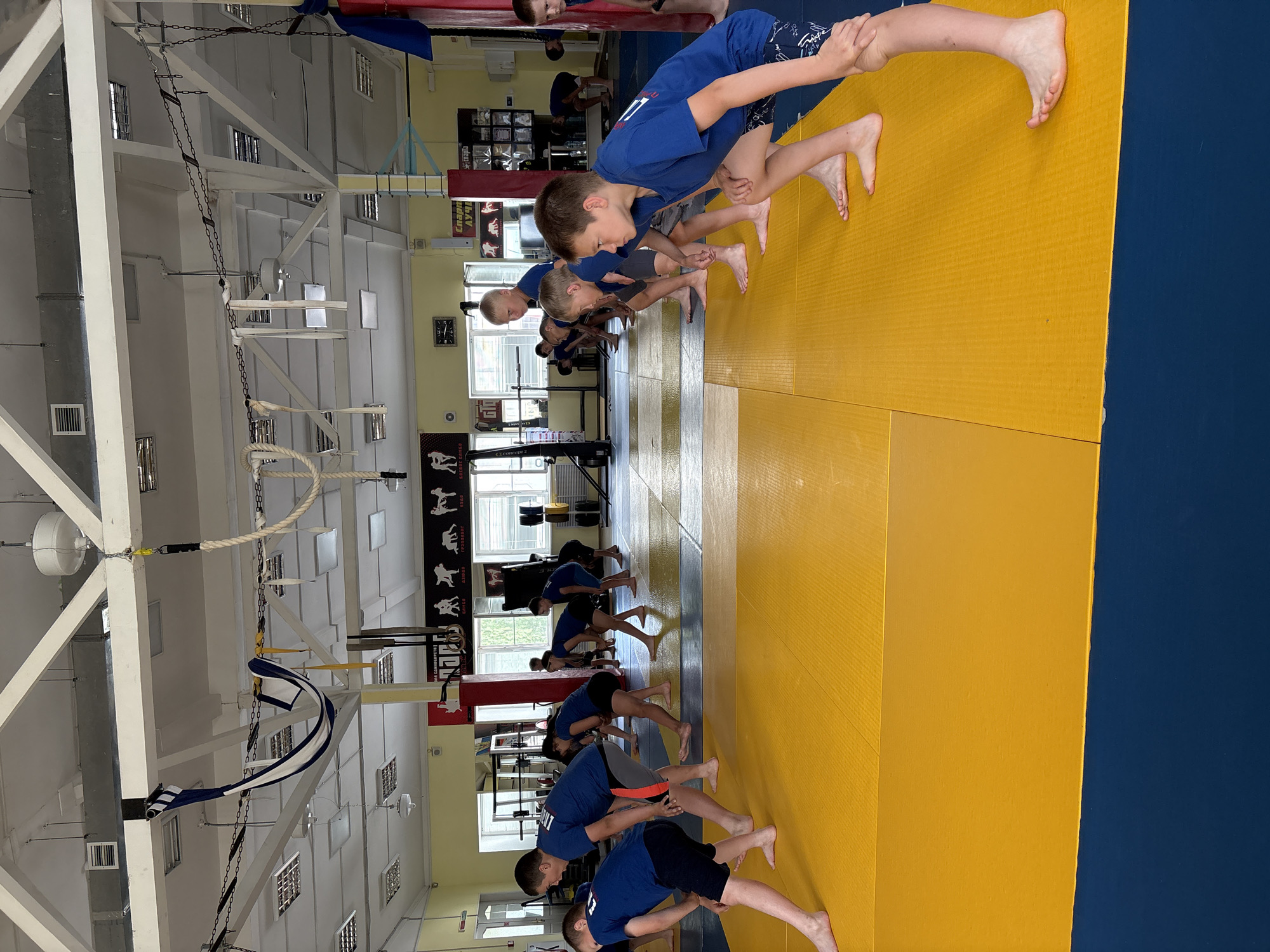 This image depicts a group of young boys in a gymnasium, likely engaged in a sports training session. They are all wearing blue t-shirts and athletic shorts. The gymnasium floor is covered with yellow and blue mats, typical for wrestling or judo training. In the background, there are gym equipment, windows, and posters on the walls, suggesting it is a dedicated sports facility. The boys are in various stages of a warm-up or exercise routine, some are in a crouching position, while others are standing and stretching. The overall scene is one of active training in an indoor sports environment. Based on the posters visible in the background with Cyrillic text, the facility is likely located in Russia. The posters feature images of athletes in various poses, and the text appears to be related to sports and potentially the specific disciplines practiced at this location. The lighting suggests it is daytime, with light coming in from the windows and artificial lights on the ceiling.