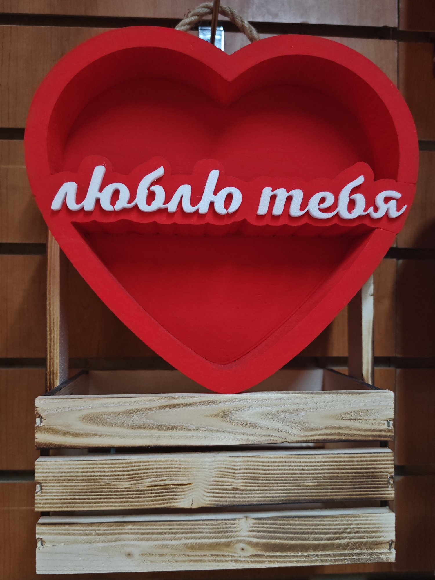 This is a vertical, eye-level shot of a decorative red heart-shaped object with white lettering on it, suspended above a wooden crate. The heart is open in the center, creating a shelf-like space. The white text on the heart is in a cursive font and reads "люблю тебя", which translates to "I love you" in Russian. The background is a wooden wall with horizontal planks. The wooden crate below the heart is made of light-colored wood with visible grain and appears to be constructed from multiple slats. The lighting is even, suggesting an indoor setting with artificial light. There are no people visible in the image, and the overall mood is decorative and potentially romantic due to the heart shape and text.