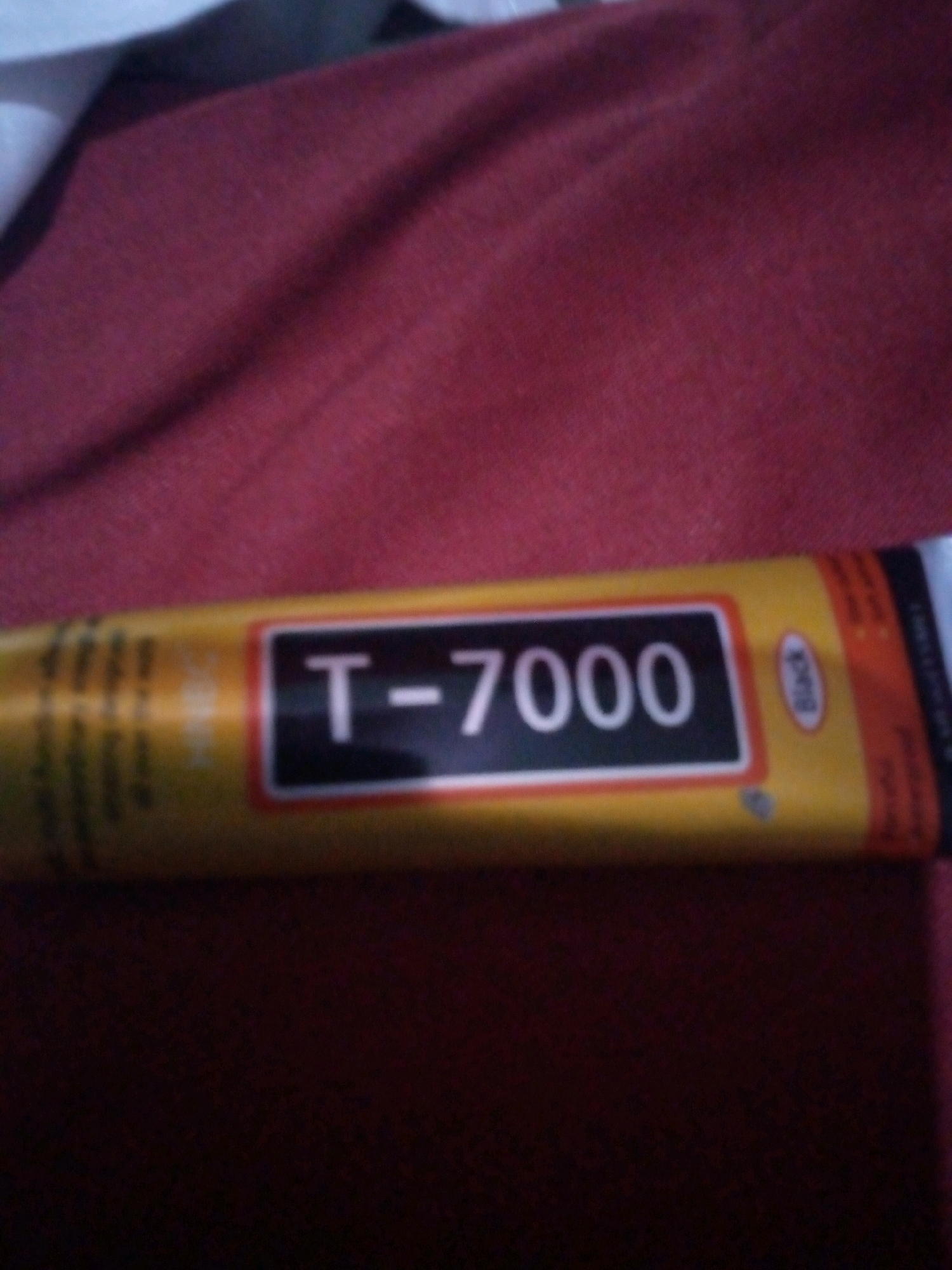 A blurry, close-up, eye-level shot shows a tube of T-7000 adhesive on a textured red surface. The tube is yellow with black and white lettering. The red surface is a textured fabric, possibly a blanket or upholstery. The image is not geographically specific, and there is no visible indication of a city or country.