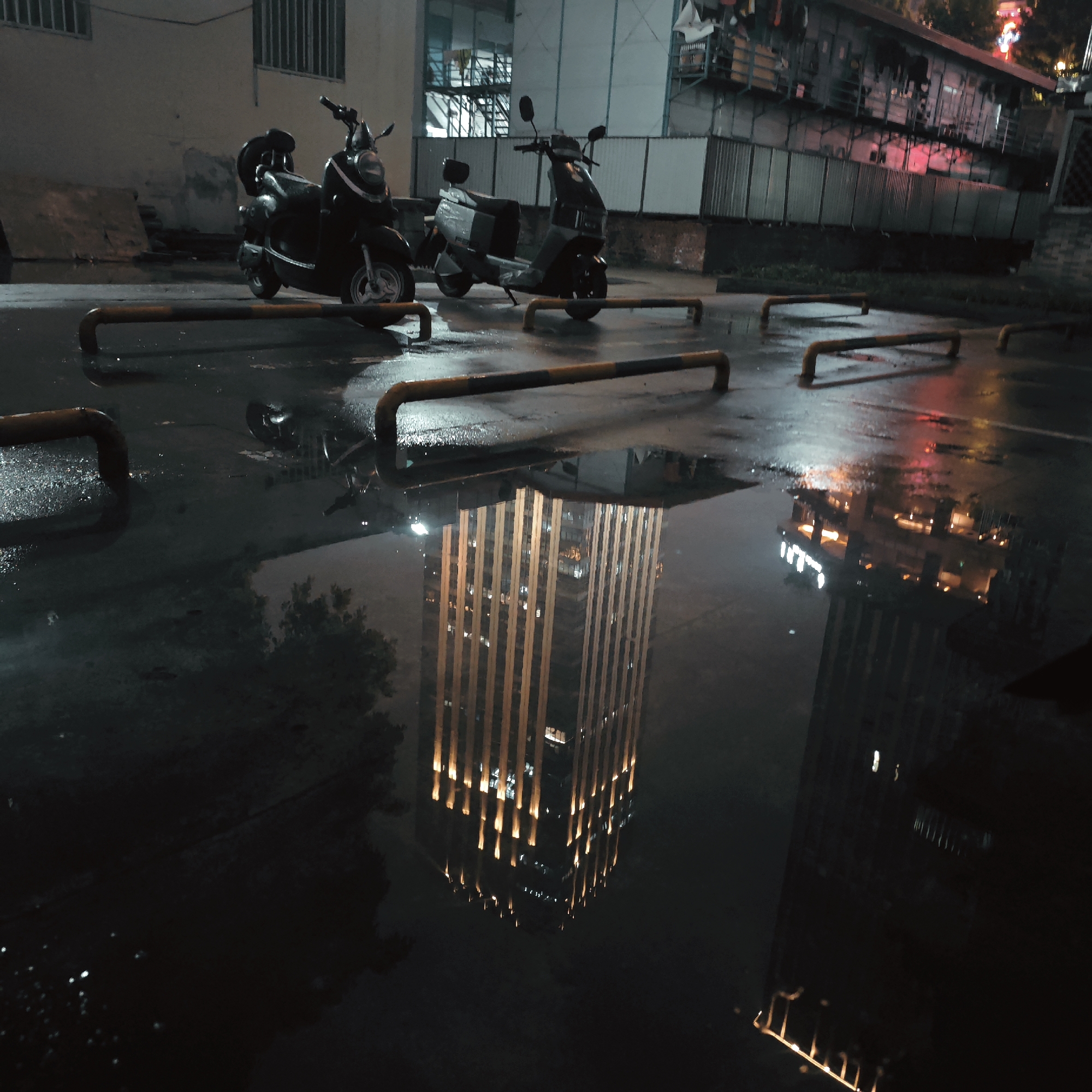 This nighttime photograph captures a quiet urban scene, likely a parking area or alleyway, immediately after rainfall, as evidenced by numerous puddles reflecting artificial lights. In the foreground and midground, two parked scooters are visible, one black and the other gray, positioned between low, yellow and black striped metal barriers.
The most striking feature of the image is the large puddle in the center, which perfectly mirrors a tall, modern building. This building's facade is dramatically illuminated by numerous vertical light strips, creating a glowing, architectural reflection that dominates the lower half of the frame. Other smaller puddles also show reflections of city lights and distant structures.
In the background, a light-colored building with barred windows is visible on the left. To the right, another structure, possibly a commercial or residential building, features faint red and white lights that also cast reflections on the wet ground. The scene is devoid of people and discernible text. The overall atmosphere is moody, serene, and atmospheric, showcasing the beauty of urban reflections at night.