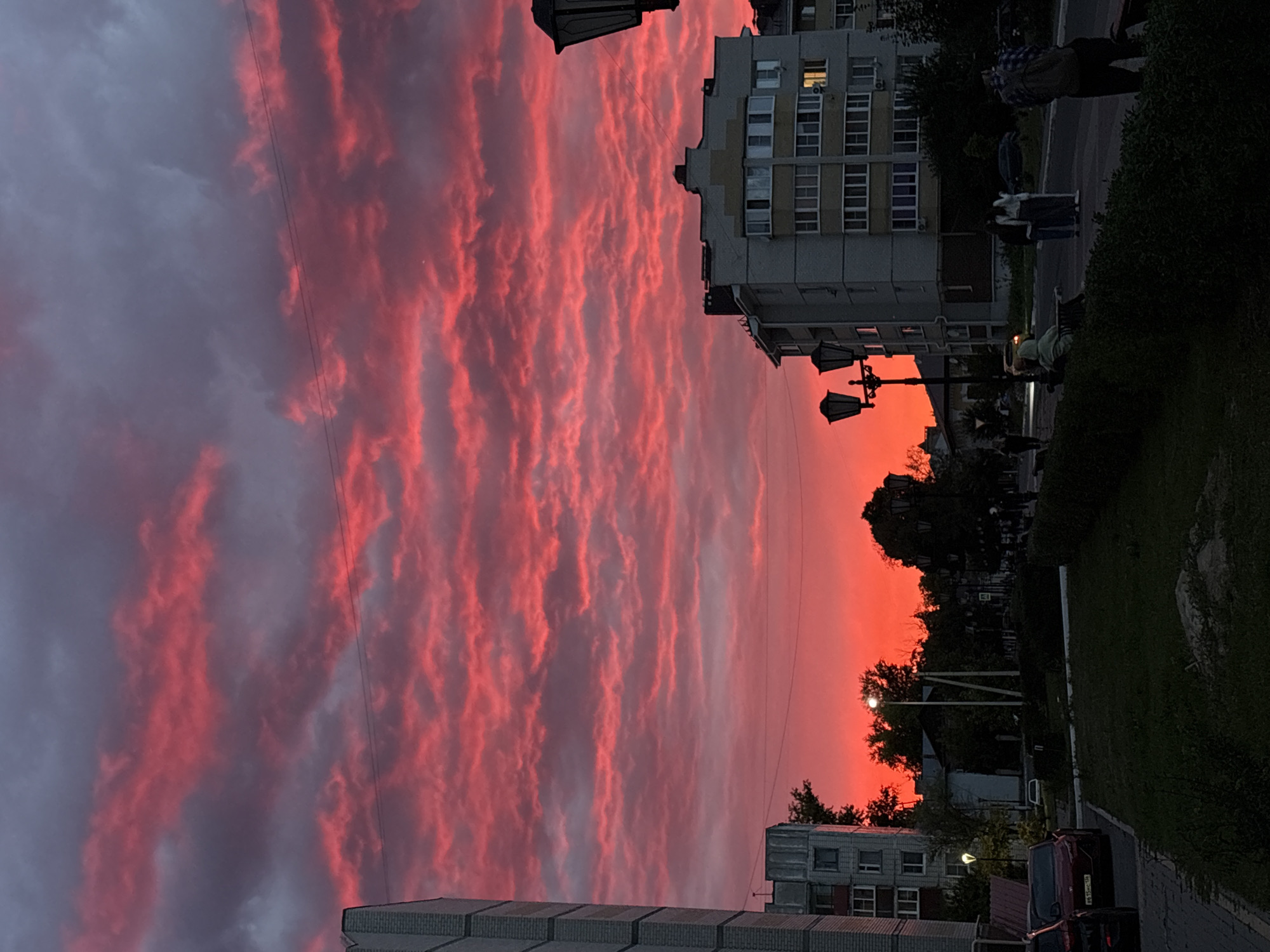A street scene in Bobrov, Russia, is depicted at dusk, with a vibrant, cloud-filled sky ablaze with crimson and pink hues. In the foreground, a grassy area with a low hedge separates the viewer from a paved sidewalk and a street. Several cars are parked along the sidewalk, including a red sedan and a black SUV.
Across the street, a multi-story residential building with numerous windows stands out. Adjacent to it are more buildings and trees, their silhouettes dark against the fiery sky. Streetlights are visible, one prominent on a pole with multiple lamps.
On the sidewalk, a few people are present. Two individuals stand together near the building, while another person sits on a bench in the foreground, facing away from the camera. A person wearing a backpack is also visible on the right side of the frame. The overall atmosphere suggests a calm evening in a residential area, with the dramatic sunset dominating the visual landscape.