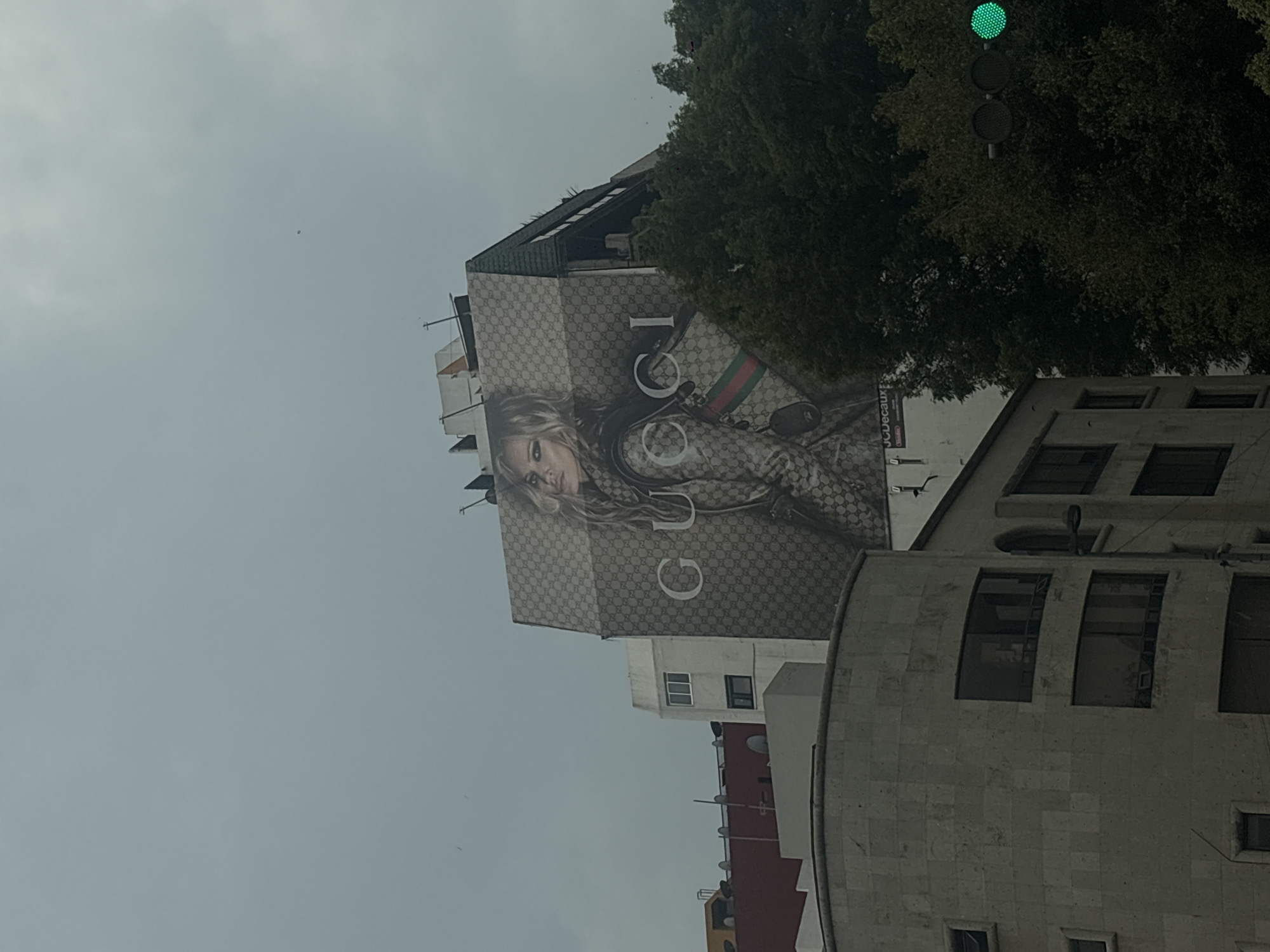 The photograph shows an exterior view of a building in Mexico City, Mexico, featuring a large Gucci advertisement spanning its facade. The ad depicts a woman and Gucci branding elements. Adjacent to the building with the advertisement is a multi-story structure with a curved facade and rectangular windows. A traffic light with a green light is visible among the trees. The sky is overcast.