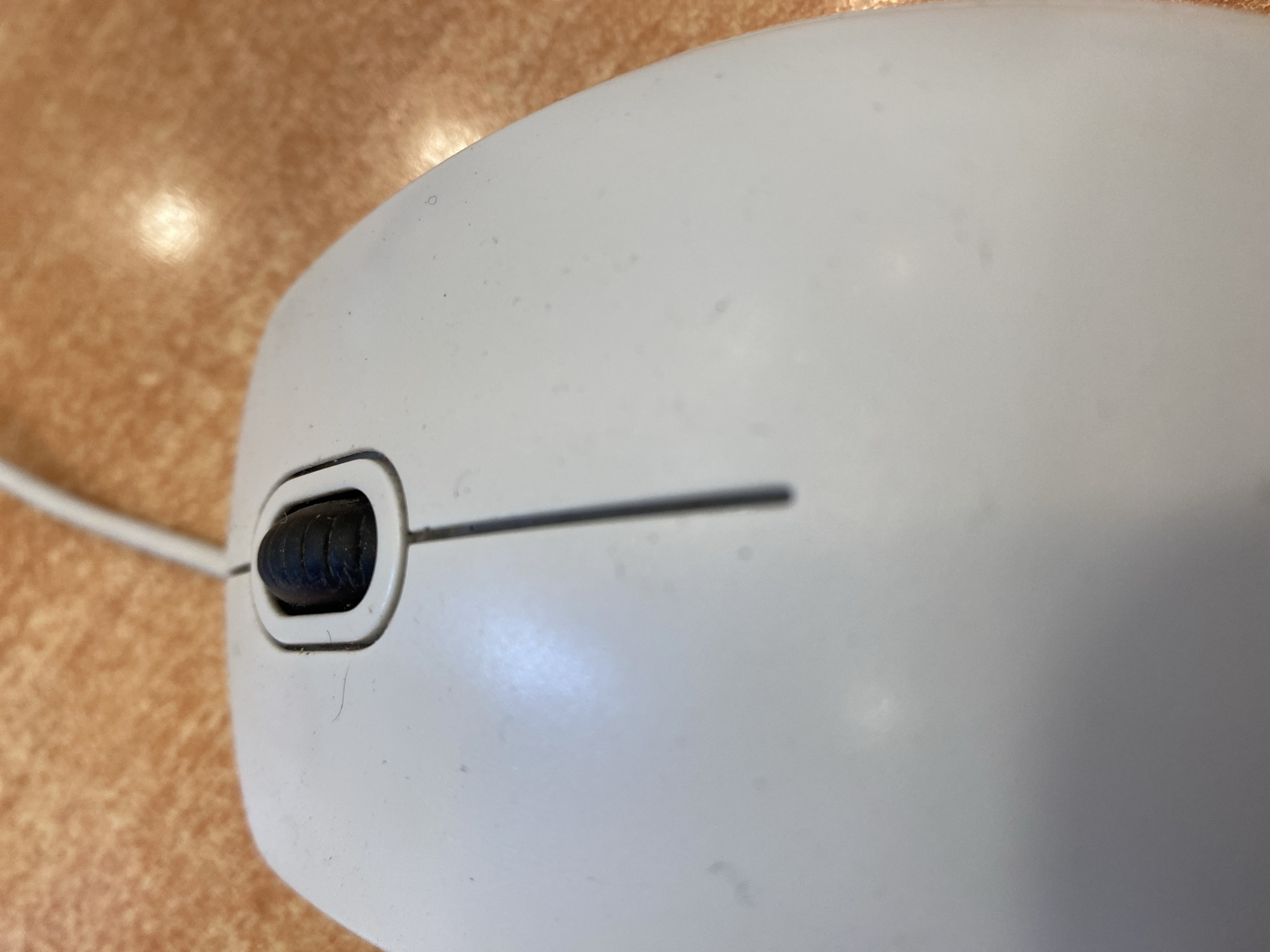 A close-up, high-angle shot captures the top of a white computer mouse resting on a textured, orange surface. The mouse has a black scroll wheel situated in the center, surrounded by a light gray plastic housing. A thin, white wire extends from the top-left of the mouse and disappears out of frame. The orange surface appears to be a table with a subtle, shiny reflection in the upper right quadrant. The scene is devoid of people or any indication of its specific location within Yerevan, Armenia.