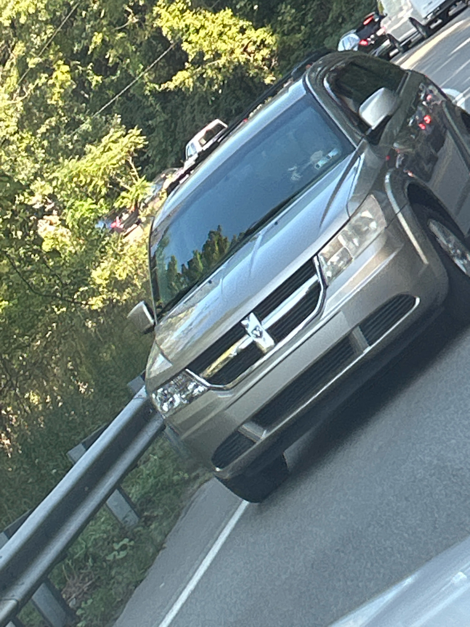A silver Dodge Journey is traveling on a road with a metal guardrail to its left and trees to the left of the guardrail. The vehicle is angled to the right, showing its front grille, headlights, and part of its windshield. The windshield reflects trees and the sky. Traffic is visible ahead of the Dodge Journey, including a white truck. The scene is likely outdoors, possibly on a highway or a suburban road, given the presence of trees and a guardrail. The image quality is somewhat grainy, and the lighting suggests it was taken during the daytime. There are no discernible people in the image. The specific location within Pittsburgh is not identifiable from the provided visual information.