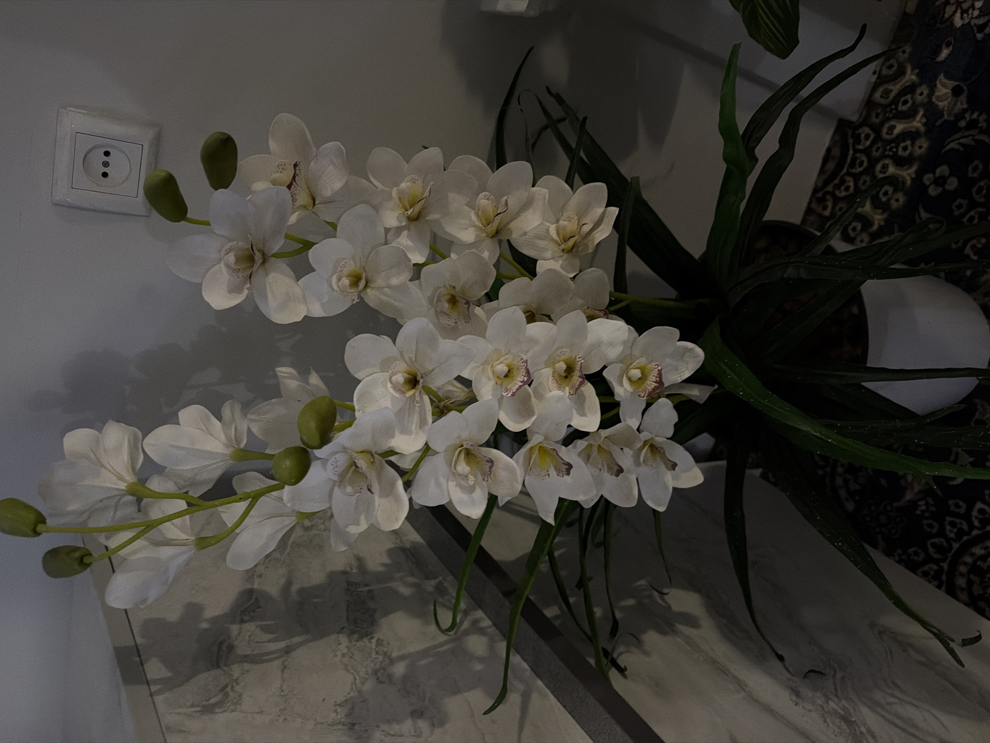 A close-up, vertical, indoor shot focuses on a faux orchid plant with multiple white blooms and green buds. The plant is in a white pot, and its long, dark green leaves drape downwards. The background is a neutral-colored wall. To the upper right of the frame, a white electrical outlet with a single socket is visible. The lower portion of the image shows a dark, patterned rug. The lighting suggests artificial light, creating shadows on the wall behind the plant. No people are present in the image. The location is identified as Khumsan, Uzbekistan.