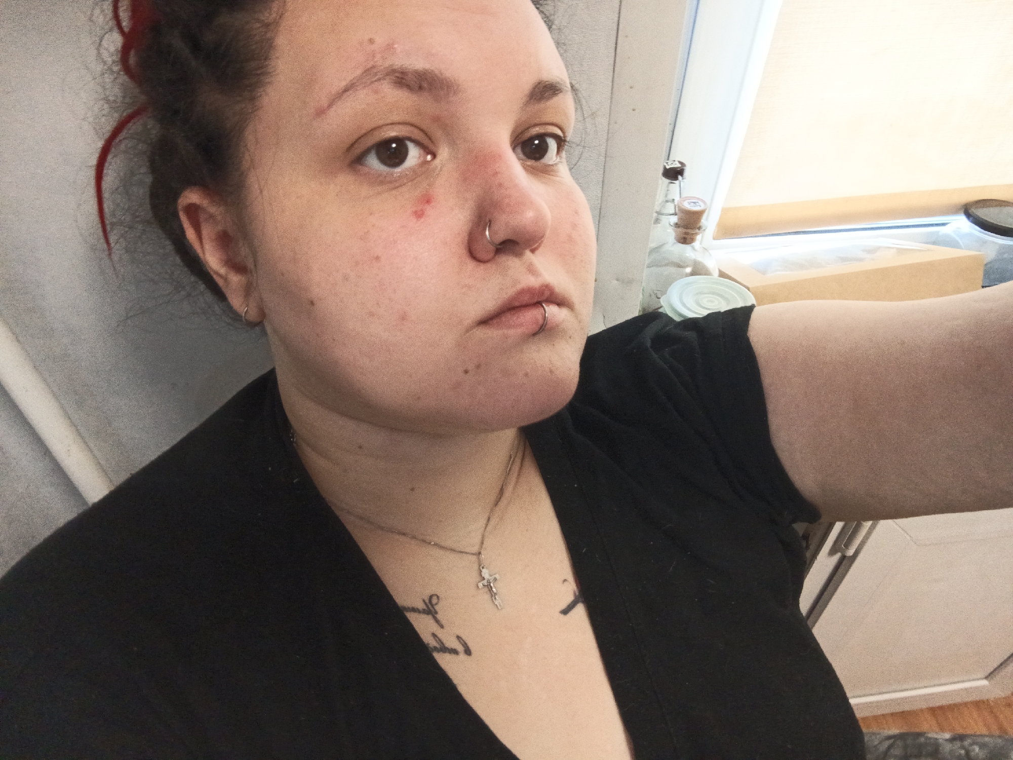 A female individual is depicted in an indoor selfie, located in the city of Chaikovsky, Russia. Her dark hair, containing visible red strands, is pulled back from her face. She has fair skin, exhibiting redness on the forehead, nose, and a distinct red lesion on her right cheek. Her nose is pierced with a silver-colored hoop, and her lower lip features two silver-colored horizontal labret piercings. A small silver-colored hoop earring is present in her left ear. She wears a black garment, a silver-colored chain necklace with a crucifix pendant, and has script-style tattoos on her chest. Her gaze is directed slightly to the right of the camera. The background on the left side consists of a light gray wall with a vertical white pipe. The right background features a window with a light-colored drawn blind, a clear glass bottle with a cork stopper, a light-colored circular lid, and a brown cardboard box with a transparent top, all situated on a windowsill or shelf. The lower right portion of the image shows a dark, textured floor surface and a light-colored cabinet or drawer unit.