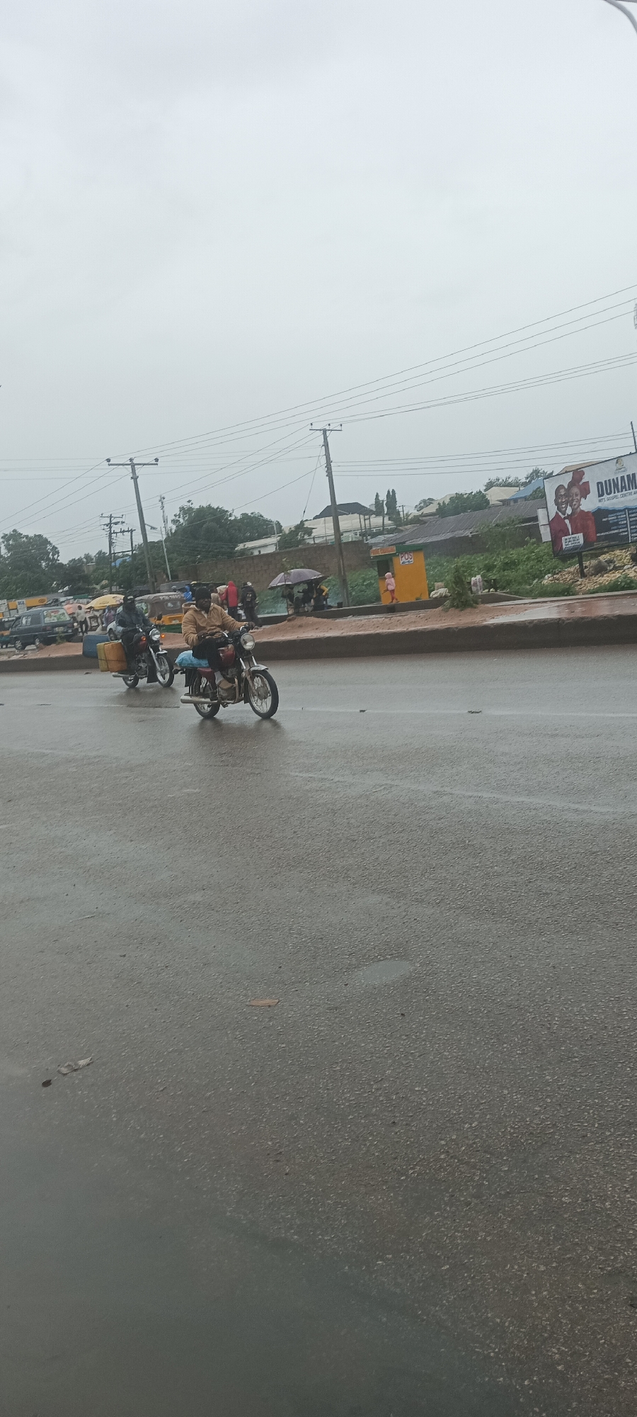 The image depicts a street scene in Bukuru, Nigeria, on an overcast day with the asphalt road visibly wet, suggesting recent rainfall or ongoing light rain. Two men are riding motorcycles on the damp road in the foreground, moving towards the right. The rider closer to the camera wears a light brown jacket, while the one behind him, in darker attire, has a large yellow container or package attached to the side of his motorcycle.
On the left side of the road, several vehicles, including cars and a motorized tricycle, are visible, along with a group of people. Many individuals in this area are holding black umbrellas, reinforcing the wet weather conditions. Utility poles and power lines crisscross the grey, cloudy sky.
On the right side of the road, a raised earth bank separates the street from the background. A small, bright yellow booth marked with red "POS" letters stands on this bank. Further back, a large billboard is visible, prominently displaying the word "DUNAMA" at the top, with smaller text beneath it including "DUNAMIS" and "INT'L GOSPEL CENTRE ABUJA," indicating an advertisement for a religious institution. The scene captures everyday urban activity and transport continuing despite the damp weather.