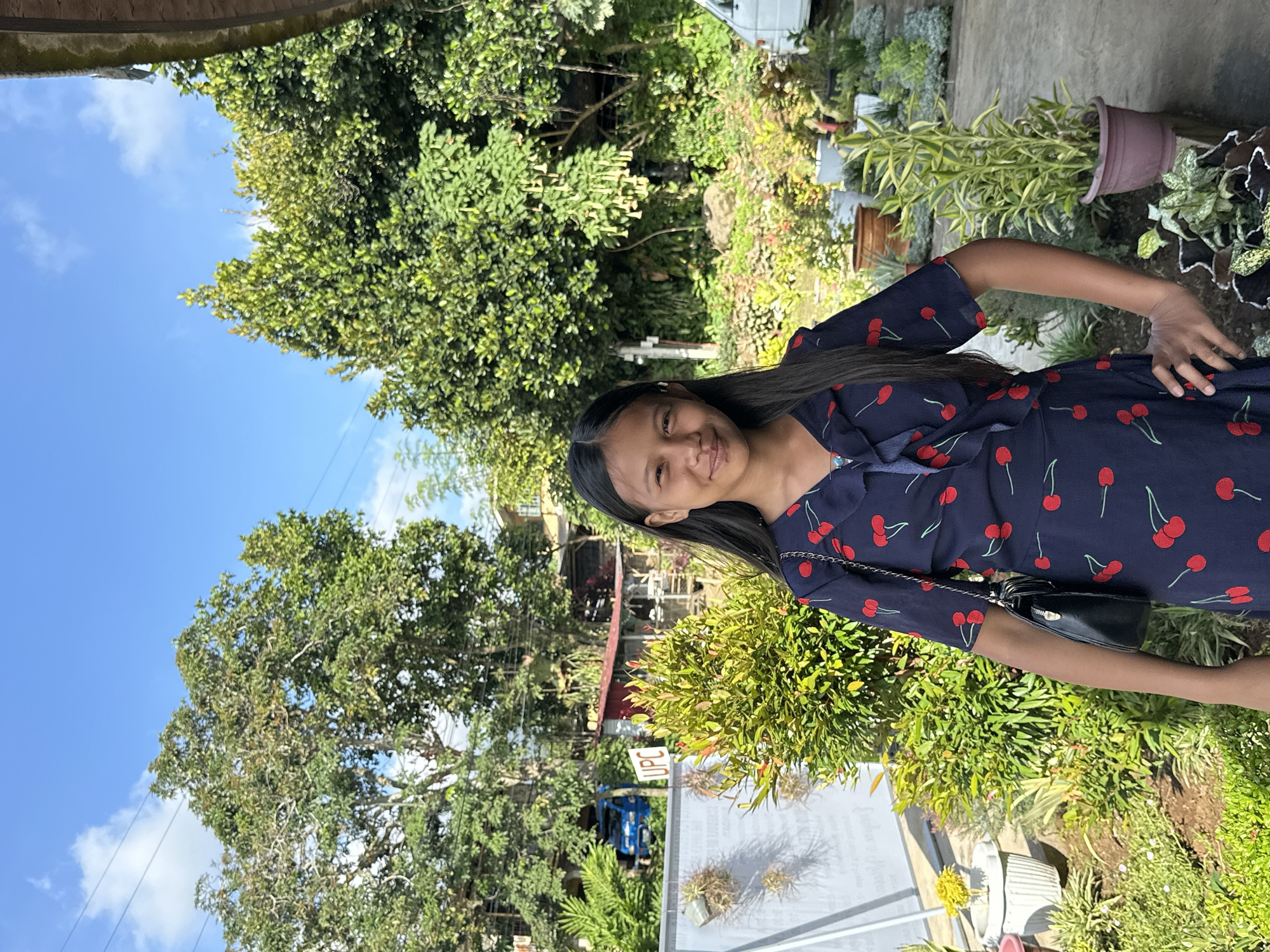 A young woman stands outside amidst lush greenery under a partially cloudy sky. She has long, dark hair and wears a navy-blue dress with a red cherry print. A black purse with a chain strap hangs on her side. The environment includes various trees and plants, some in pots. A sign with "UPC" is visible in the background. The scene is outdoors, with glimpses of buildings and a vehicle, suggesting an urban or suburban setting. Given the prompt, the location is Davao City, Philippines.