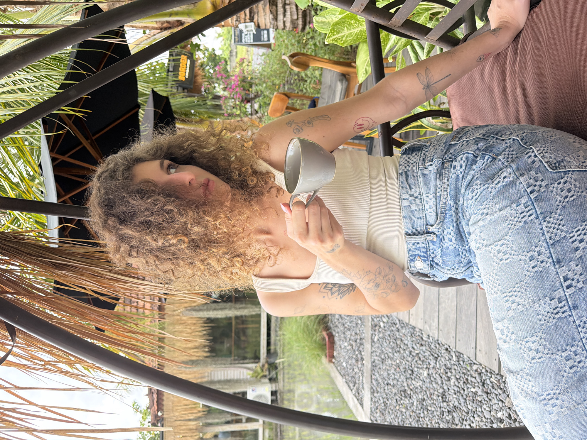 A medium shot captures a young woman with curly brown hair sitting on a dark brown cushioned swing seat. She is wearing a white ribbed tank top and light blue patterned jeans. The woman is holding a small gray mug in her right hand and looking to her left. Her left arm rests on the seat, revealing several tattoos. She has a large floral tattoo on her forearm and a dragonfly tattoo on her upper arm.
The environment suggests an outdoor cafe setting in Mengwi, Indonesia. In the background, there is a body of water with gravel paths and lush green vegetation. Wooden walkways and outdoor seating are visible, along with thatched roof structures. Two black umbrellas are open, and a sign for "BORDER COFFEE & GREEN" is partially visible. Palm fronds and other tropical plants frame the scene, indicating a warm climate. The overall atmosphere is relaxed and casual.