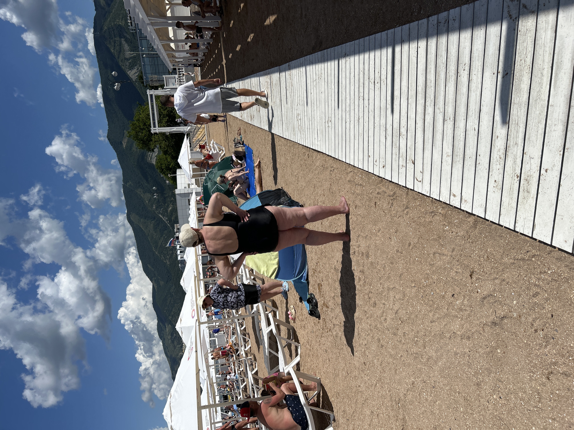 The image depicts a beach scene in Gelendzhik, Russia, under a bright, cloudy sky. In the foreground, a woman in a black swimsuit and a light-colored hat stands facing away from the camera, her hands on her hips. To her left, a man in a patterned shirt and shorts is seated on a lounge chair, looking towards a child. A light blue towel and a pair of black sandals are on the sand near the woman.
Further down the beach, a white wooden boardwalk extends into the distance. A man in a white hoodie and grey shorts walks away from the camera along the boardwalk. Several white beach umbrellas and lounge chairs are set up on the sand, with various people relaxing and sunbathing. In the background, a line of white-covered structures, likely beach cabanas or cafes, runs parallel to the shore. Beyond the structures, lush green mountains rise under the expansive sky. The overall impression is a typical summer day at a popular seaside resort.