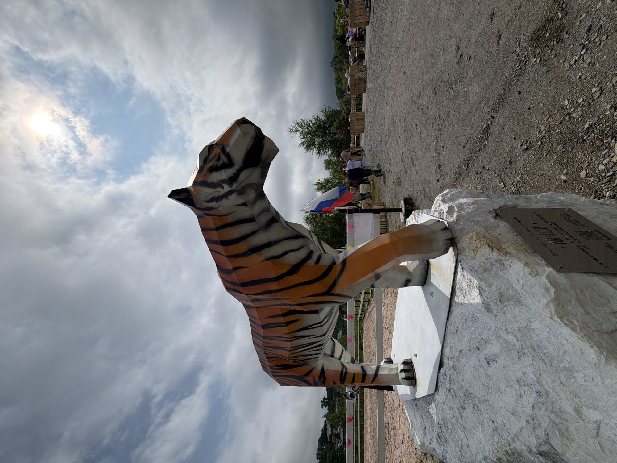 A low-angle, wide shot captures a polygonal sculpture of a tiger standing on a large, light-colored rock. The tiger sculpture is orange with black stripes and appears to be made of metal or plastic. The rock it stands on has a metal plaque attached to its base. In the background, a dirt path leads to a fenced-off area with wooden crates and tables set up, suggesting an outdoor event or market. Several people are visible in the background, some walking and others standing near the tables. A Russian flag is planted to the left of the tiger sculpture. The sky is overcast with patches of sunlight breaking through, indicating a partly cloudy day. The environment suggests an outdoor location in a town or rural area. The plaque on the rock indicates this sculpture is located in Teploozersk, Russia, and was installed on June 20, 2023.