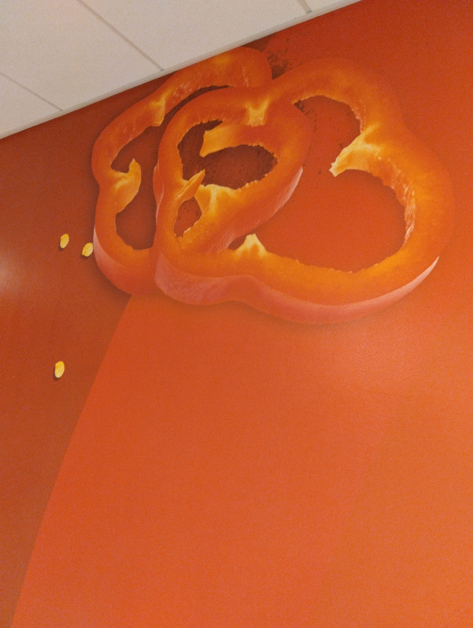 The image is a close-up, slightly angled shot of a mural or wall art depicting two slices of bell pepper. The bell pepper slices are a vibrant orange-red and are rendered with a glossy finish, suggesting they are painted or printed onto the surface. The slices are positioned in the upper portion of the image, overlapping each other. Several small, pale yellow bell pepper seeds are scattered on the surface below the pepper slices. The background is a solid, matte orange color, with a subtle gradient that creates a sense of depth. The upper edge of the image shows a sliver of a white ceiling with linear texture. The scene is indoors, and there are no people or animals visible. The lighting is soft and diffused, highlighting the texture of the orange background and the shine of the bell pepper artwork. The location is not specified within the image content.
