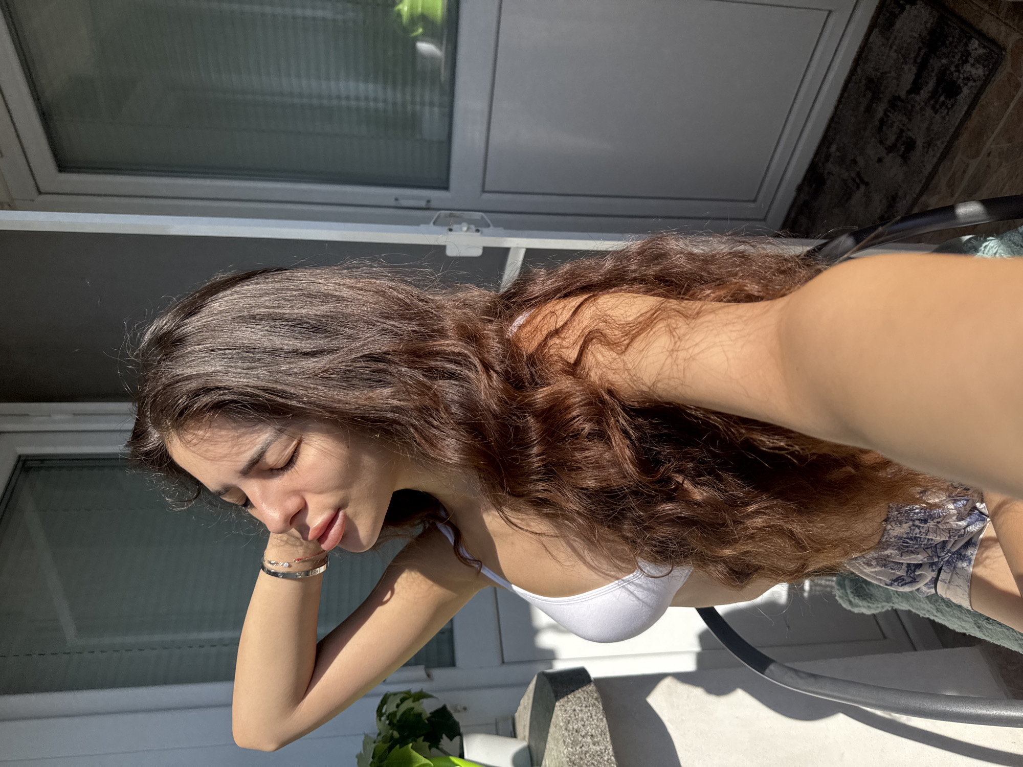 A young woman with long, wavy brown hair is sitting on a chair on a balcony. She is wearing a white bikini top and patterned shorts. Her eyes are closed and her head is tilted back, with one arm raised behind her head. The sun is shining brightly, casting shadows on the balcony. In the background, there is a glass door with blinds and a plant on a ledge. The scene suggests a relaxed and sunny atmosphere on a balcony in Plovdiv, Bulgaria.
