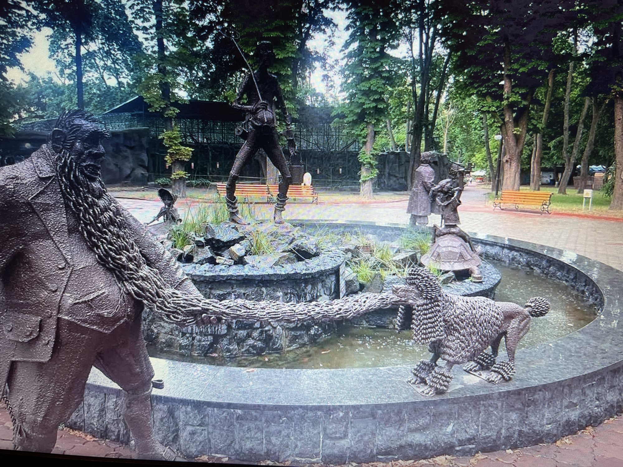 In Bilenke, Ukraine, a bronze fountain depicts a series of figures interacting around a tiered water feature. In the foreground left, a large, bearded man in a suit and breeches is connected by a thick, braided chain to a bronze poodle on the right. The poodle stands on the edge of the fountain's basin. In the center of the fountain, a figure resembling a hunter or fisherman, with a fishing rod over its shoulder and a bucket, is posed in a dynamic stance. Behind the hunter, other bronze figures are visible, including two individuals who appear to be a man and woman, and a turtle. The fountain is surrounded by a paved walkway and set within a park-like environment with mature trees and benches. The water in the fountain is murky with fallen leaves visible.