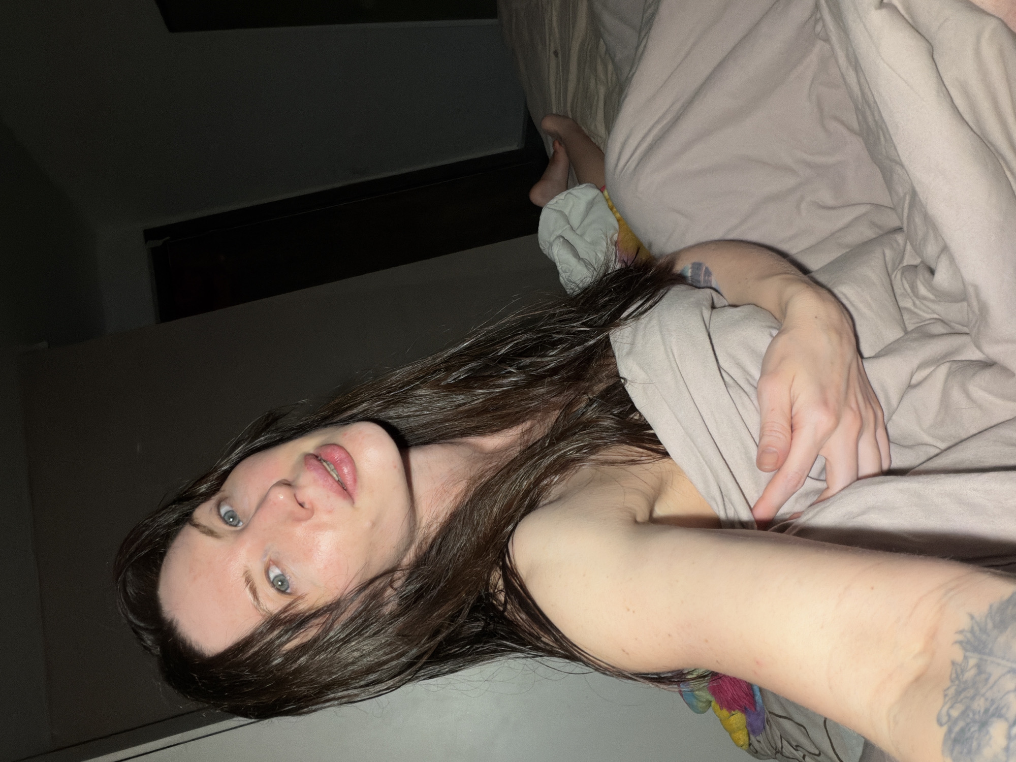 A woman lies in bed under beige bedding. She has long, dark, wet hair and blue eyes. A tattoo is visible on her left arm. The environment is dimly lit. No information suggests the image was taken in Perm, Russia.