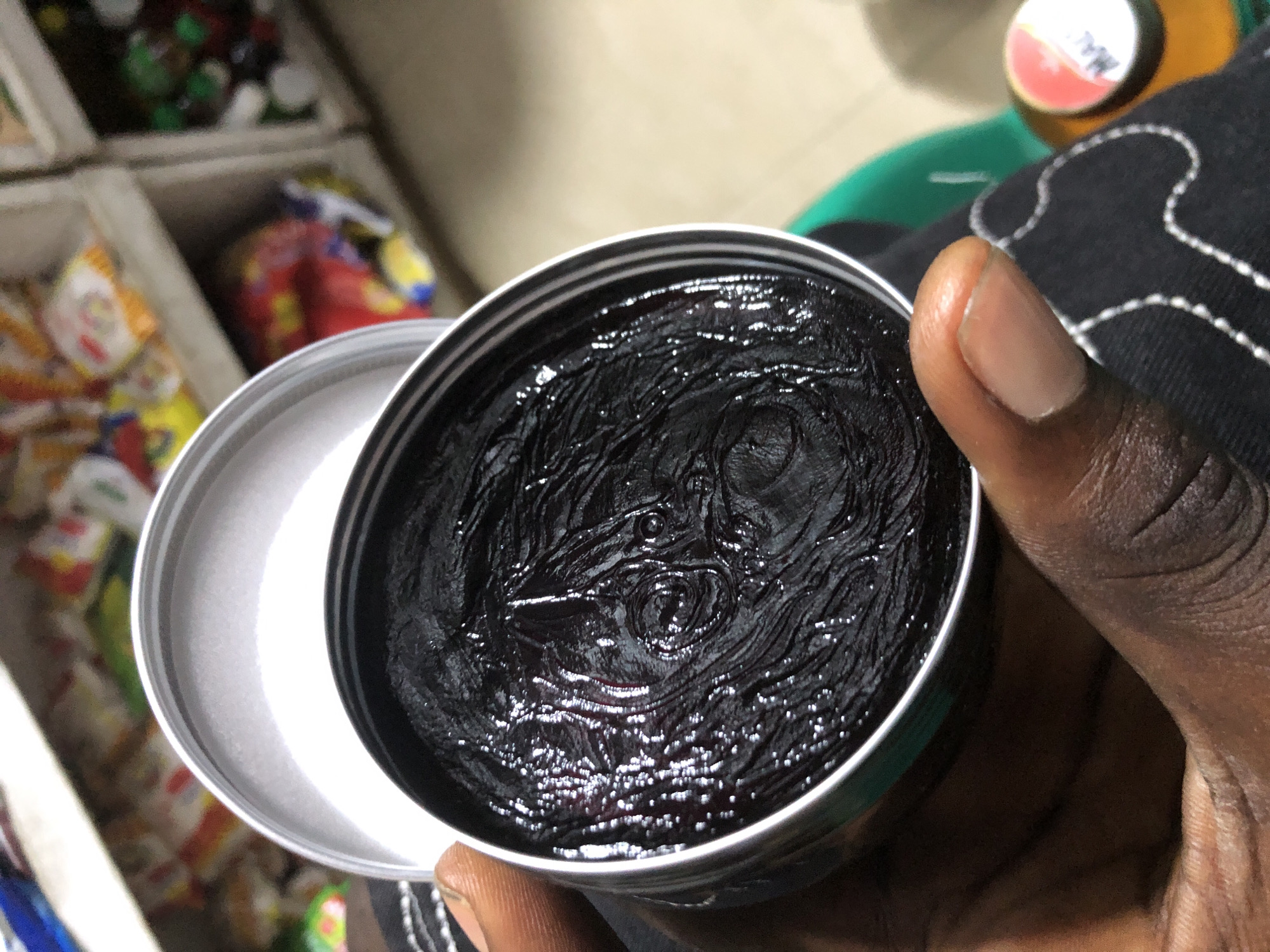 The image shows a close-up of a hand holding an open tin of dark, viscous product, likely shea butter or a similar cosmetic item, in Ado, Nigeria. The product has a textured surface with visible swirls and some lighter, shiny specks. The tin is metallic and slightly reflective. The background is blurred but suggests a retail environment with shelves stocked with various packaged goods, some appearing to be food items or drinks. A portion of a black garment with white stitching is visible in the foreground, and a bottle cap with a red and white logo is partially visible to the right. The lighting is somewhat dim, with reflections on the product and tin indicating an indoor setting.