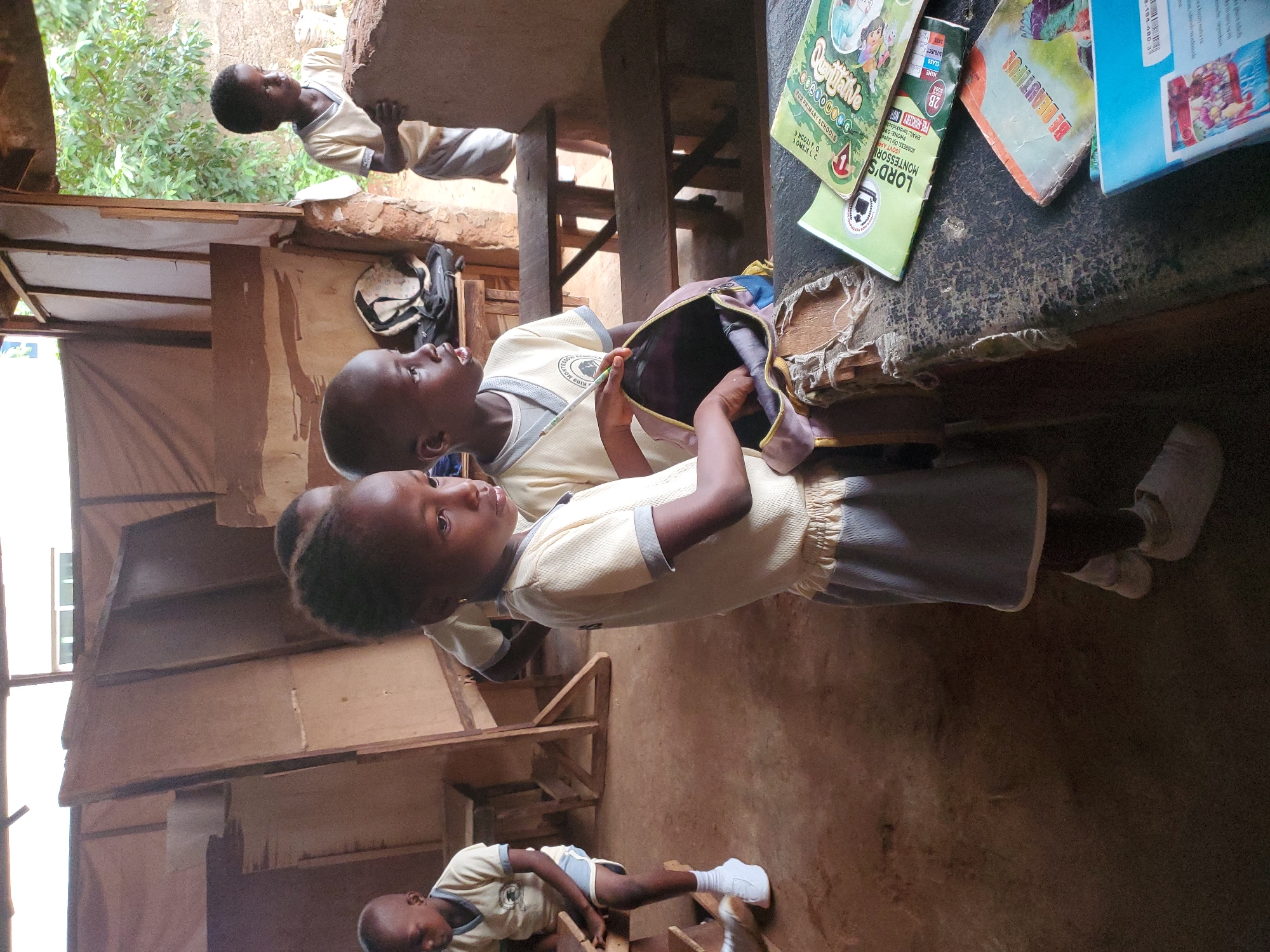 The image shows several school children in a classroom setting, likely in Ukwu-Abwa, Nigeria. Two children are prominently positioned near a table with schoolbooks, including one titled "Quittalive" and another from "Lord's Montessori". One child is looking into an open school bag. Other children are seen in the background, seated on what appear to be wooden benches or chairs. The classroom appears simple, with wooden walls and a dirt floor.