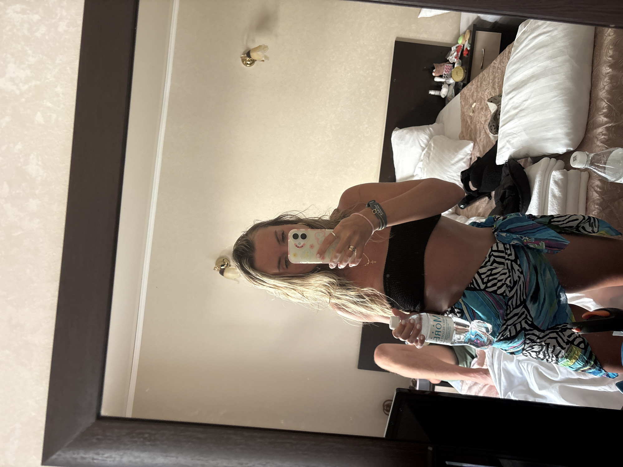 This media file shows a reflection in a mirror of a young woman taking a selfie. She is wearing a black bikini top and a colorful sarong tied around her waist. The woman has long, blonde, wavy hair and is wearing jewelry, including rings and bracelets. She is holding a smartphone in her left hand and a bottle of water in her right. The background of the reflection reveals a hotel room, with a bed, pillows, and a nightstand with various items on it. The overall lighting suggests it is daytime, and the setting appears to be a domestic or hotel interior. The woman's expression is neutral, and the scene is casual. Text visible on the water bottle is "MORE" in a stylized font.