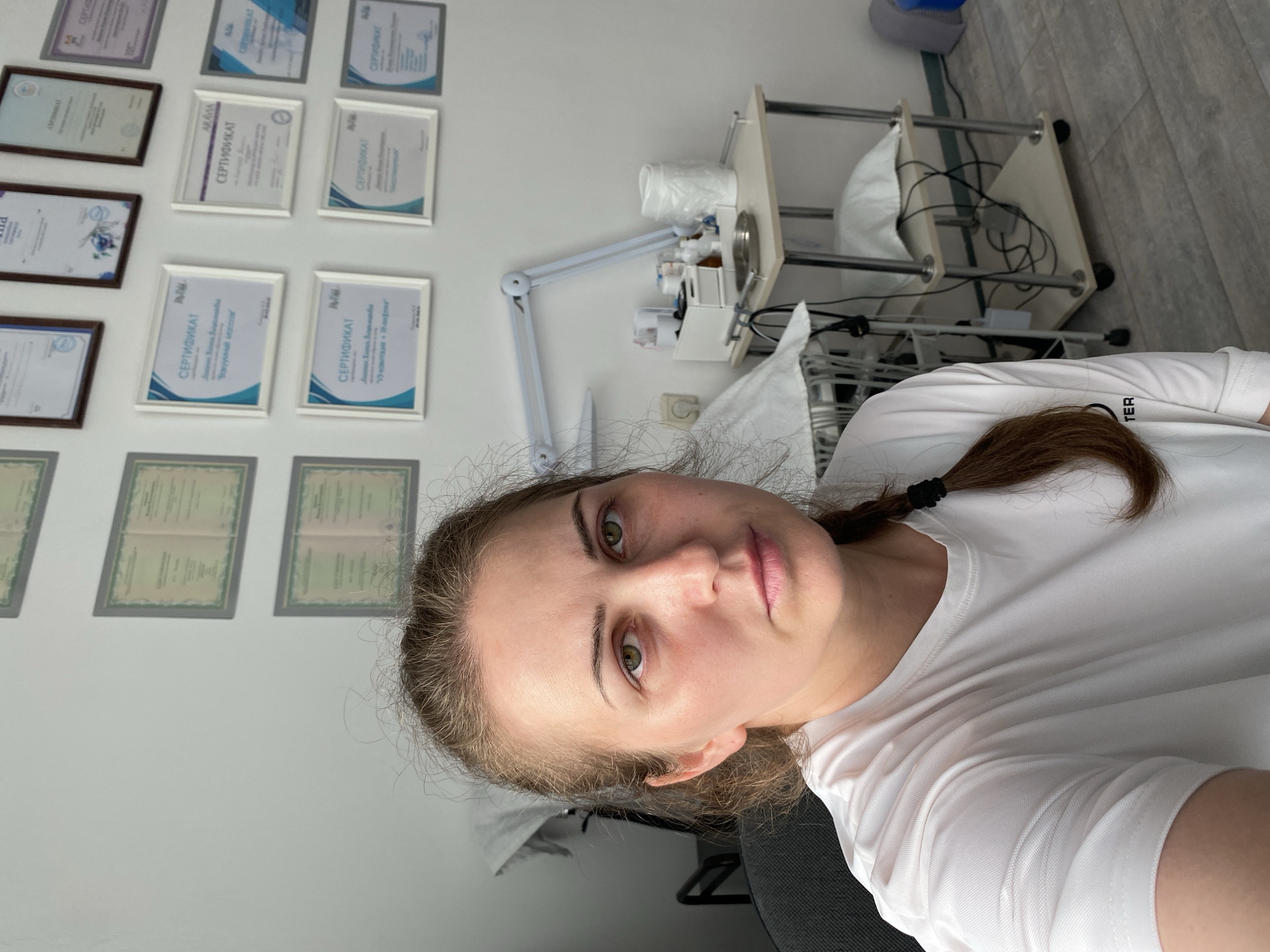 The image depicts a woman in a close-up selfie, seated in what appears to be a professional or clinical setting. She has light skin, green eyes, and brown hair pulled back in a braid. She is wearing a white t-shirt. Behind her, the wall is adorned with several framed certificates or diplomas. To the right, there is a medical or treatment table with various bottles and supplies, along with a lamp. The overall environment suggests a beauty salon, a medical office, or a similar establishment. The location provided, Марковский, Russia, contextualizes the setting within that specific geographic area.