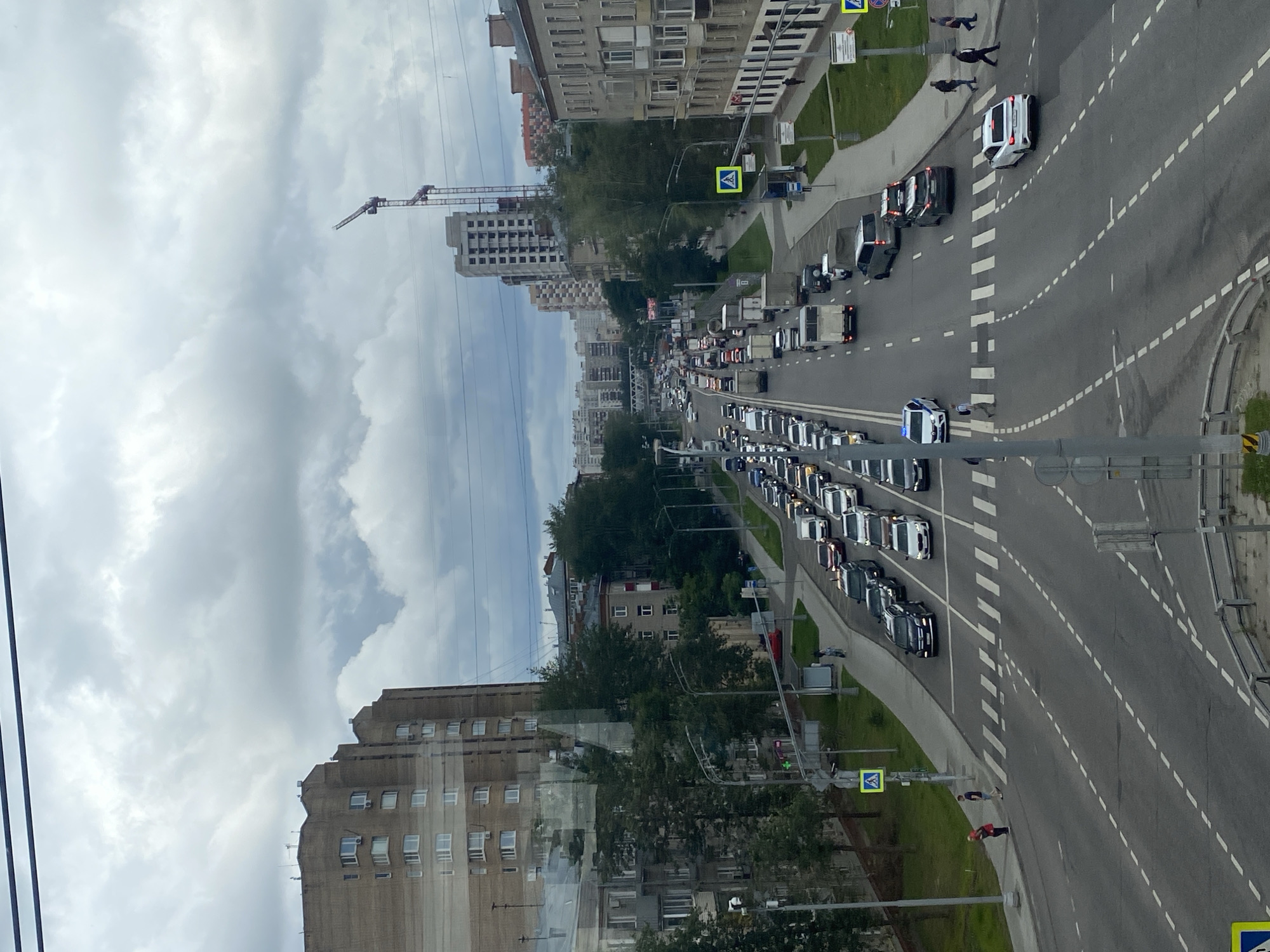 This image depicts a wide, multi-lane road in Moscow, Russia, choked with traffic. The perspective is from an elevated viewpoint, looking down at the congested street. On the left, a tall, Brutalist-style brick apartment building dominates the foreground, its facade punctuated by numerous windows. Further down the street, more residential buildings and some trees are visible.
The road itself is filled with a long queue of vehicles, including cars, trucks, and vans. Pedestrians can be seen on the sidewalks and crossing the street at marked crosswalks. Several traffic lights are visible, some of which appear to be red, contributing to the traffic jam. A construction crane is visible in the distance, suggesting ongoing development in the city. The sky is overcast with heavy clouds, casting a muted light over the scene. The overall impression is one of a busy, densely populated urban environment during daylight hours.