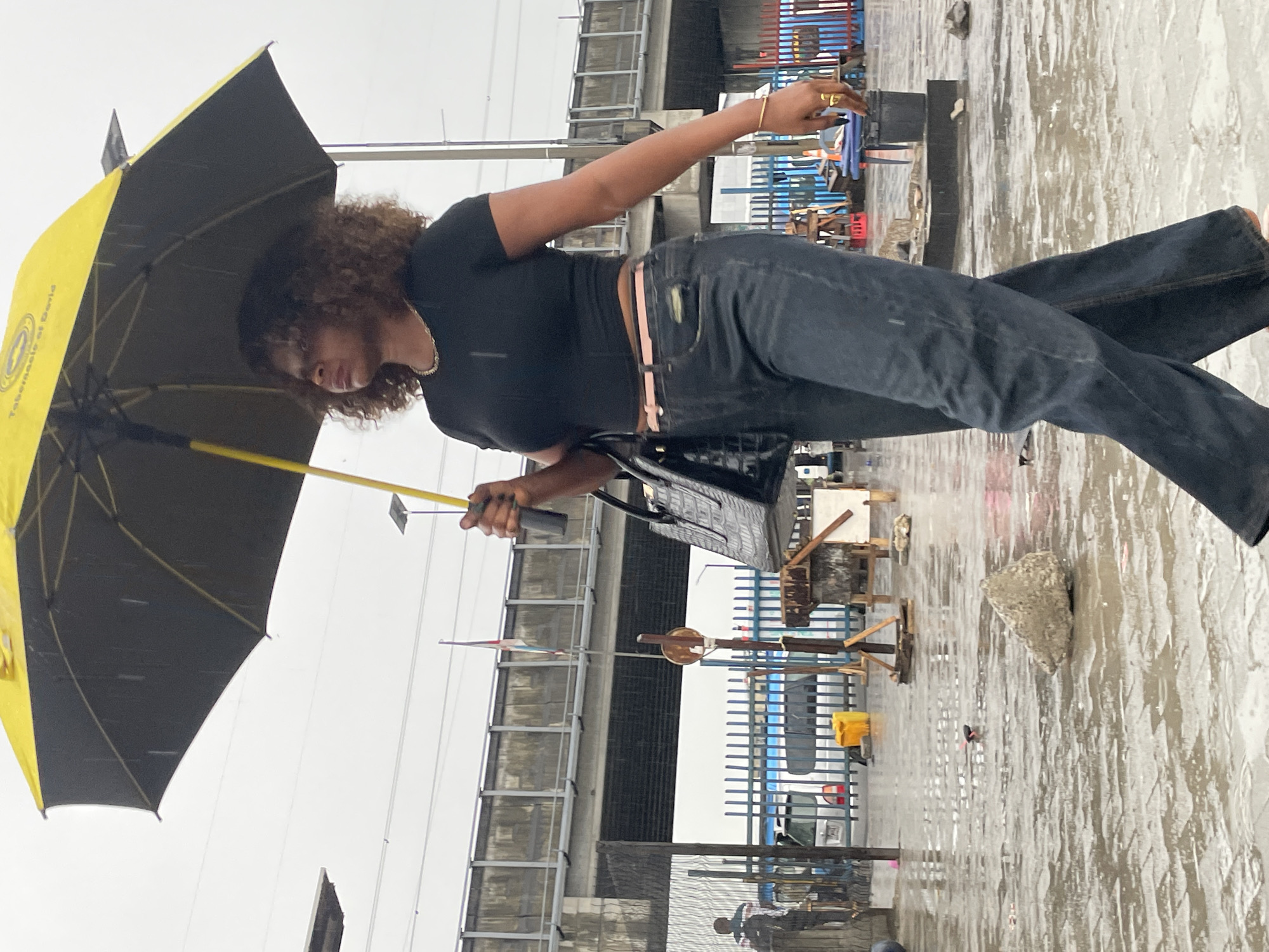 A woman stands holding a black and yellow umbrella in Lagos, Nigeria during rainfall. She is dressed in a black shirt, jeans, and a pink belt. The ground is flooded and there are various structures and items scattered around, including a fence, vehicles, and pieces of wood. The setting appears to be a commercial or market area with overcast skies.