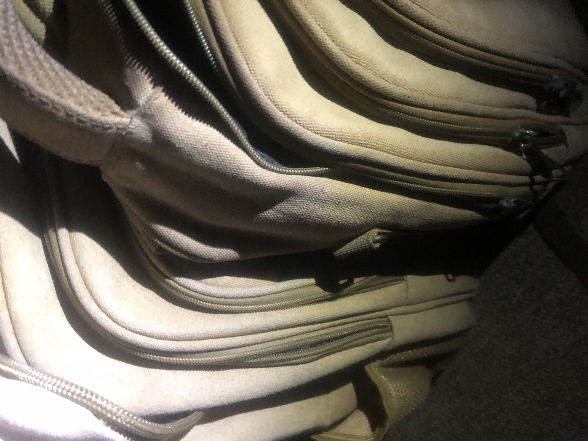 The image shows a close-up, angled view of multiple stacked zippers on tan-colored fabric. The zippers are aligned closely together, creating a series of parallel lines and curves. The fabric appears to be textured, possibly canvas or a similar material. Shadows cast by the zippers and fabric create a dramatic play of light and dark across the surface. There is no identifiable urban or natural landscape visible, making it impossible to determine a specific location within Jalingo, Nigeria, or any other city. The scene is static and devoid of any human presence or activity.