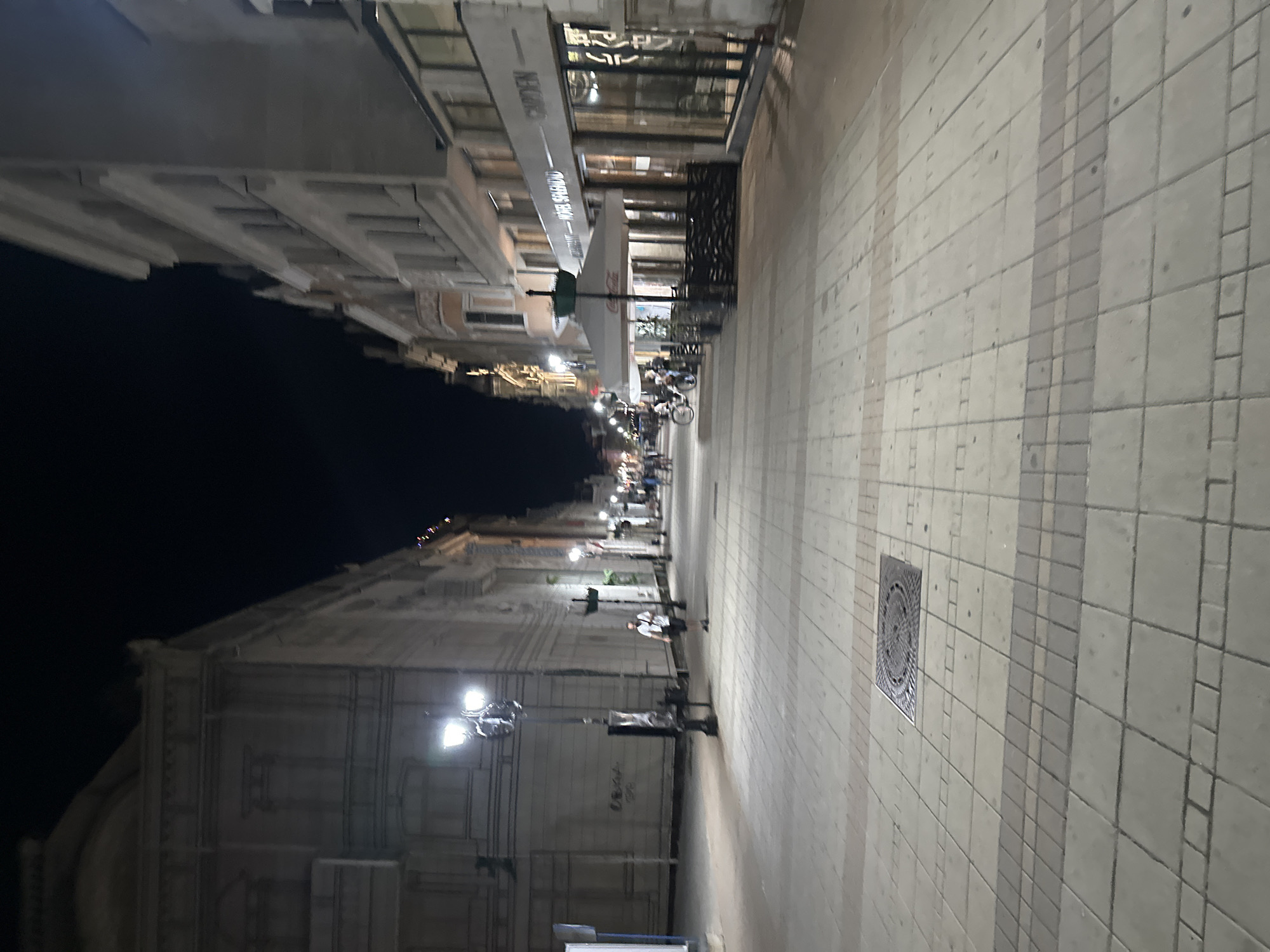 The image depicts a nighttime view of a pedestrian street in Ruse, Bulgaria, likely Alexandrovska Street, a prominent commercial and pedestrian zone. On the left, a large, ornate building with classical architectural elements is visible, possibly the Ruse Opera House or a similar cultural institution. Streetlights illuminate the broad, paved walkway, which is laid with rectangular stone tiles.
To the right, a row of buildings with shops and cafes lines the street. One establishment, labeled "RESTAURANT - HOTEL SPLENDID," features an outdoor seating area under a white umbrella canopy with a Coca-Cola logo. Another sign reads "CAMDEN." The buildings have multiple stories with visible windows.
Several people are present on the street. A lone man in a white t-shirt and dark shorts walks away from the camera. In the distance, a few individuals can be seen near the outdoor seating area, and a person is riding a bicycle. The overall atmosphere is calm and illuminated by artificial lighting, suggesting it is evening or night. The street is clean and well-maintained, with a manhole cover visible in the foreground.