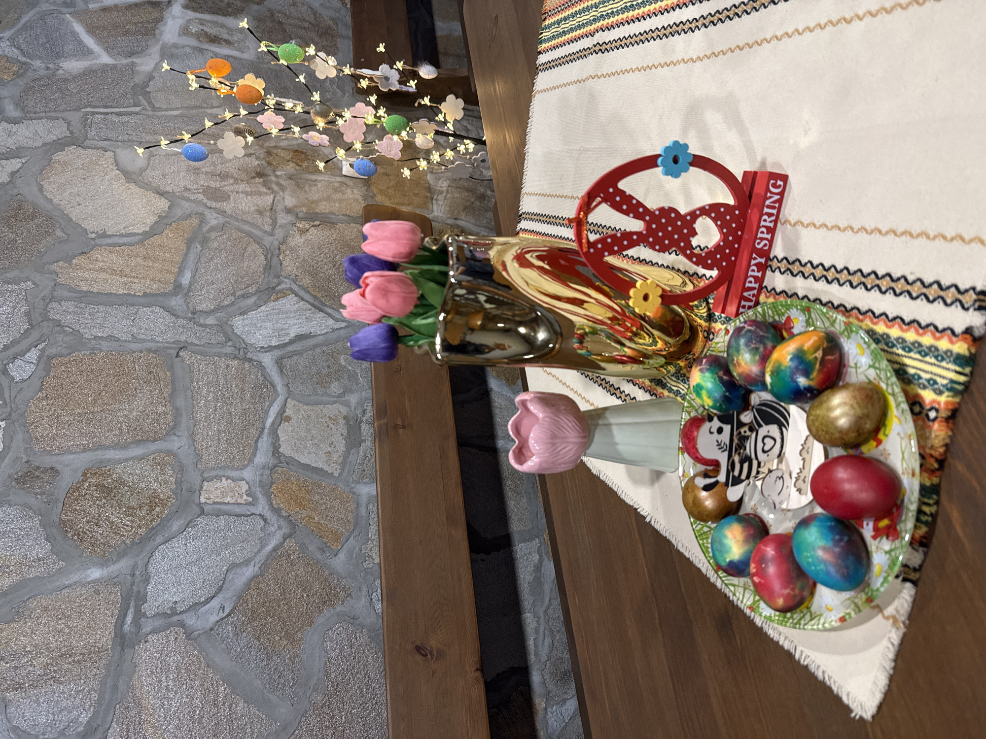 The image depicts an Easter display, likely indoors. There is a stone wall and wooden table. A bouquet of pink and purple tulips is in a gold vase and a ceramic tulip vase sits to the side. On the table is a decorative tablecloth with a geometric pattern. A plate of dyed Easter eggs sits on the table. There is a "Happy Spring" decoration next to the eggs. An Easter tree decorated with lights, flowers and eggs is placed in the background. While the location is stated to be Lukovit, Bulgaria, there is nothing in the image to confirm or deny this.