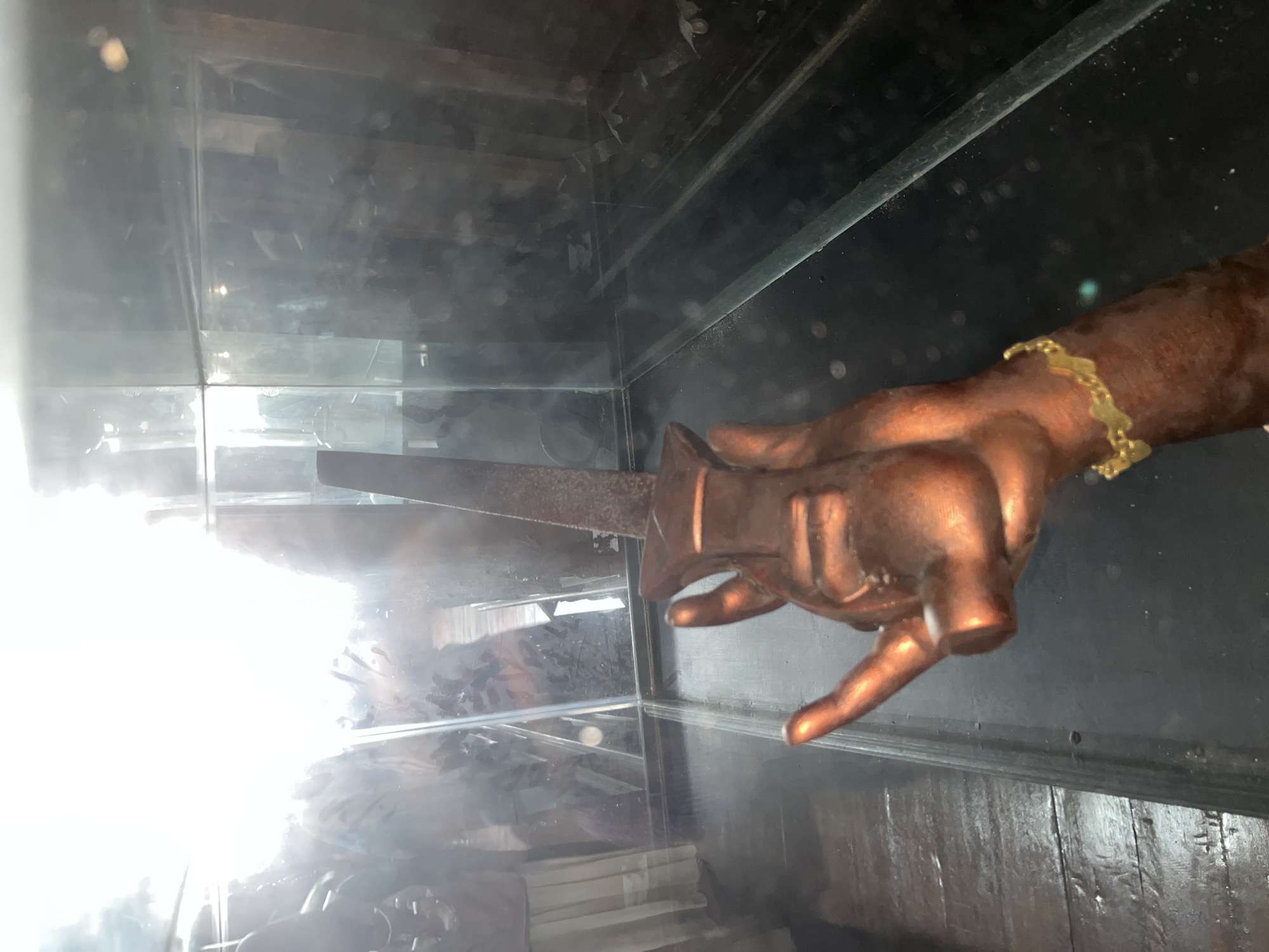 A copper-colored hand sculpture, holding a metallic dagger, is positioned in the foreground. The sculpture is within a glass enclosure that reflects its surroundings, including what appears to be a wooden floor and veiled figures in the background. A gold bracelet adorns the wrist of the sculpture. The scene is brightly lit from the left, causing significant glare and obscuring details in that area. The location is described as Mesjidpirak, Indonesia.