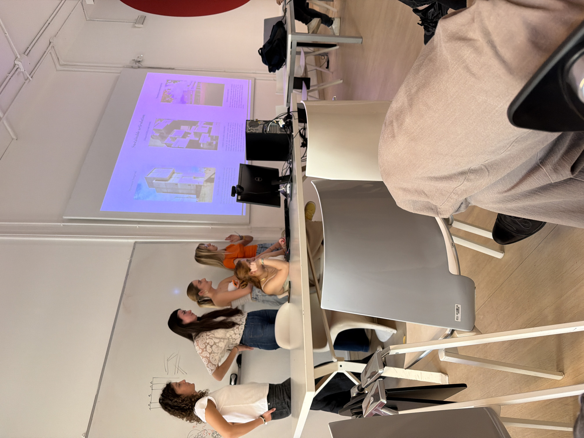 A medium shot shows a classroom scene at an educational institution in Milan, Italy. Four young women are standing in a line in front of a whiteboard, looking towards a projector screen displaying architectural renderings. From left to right, the first woman has curly brown hair, wearing a white shirt and black pants. The second woman has dark hair, wearing a white lace shirt and jeans. The third woman has light brown hair, wearing a cream-colored top and jeans. The fourth woman has long blonde hair, wearing an orange top and jeans. In front of the women is a long white table with chairs. On the table are computer monitors and components. The whiteboard has some sketches on it. The projector screen is displaying images of what appears to be building designs. In the foreground, the side of a person wearing beige pants and black shoes is visible. The classroom is well-lit and appears to be modern.