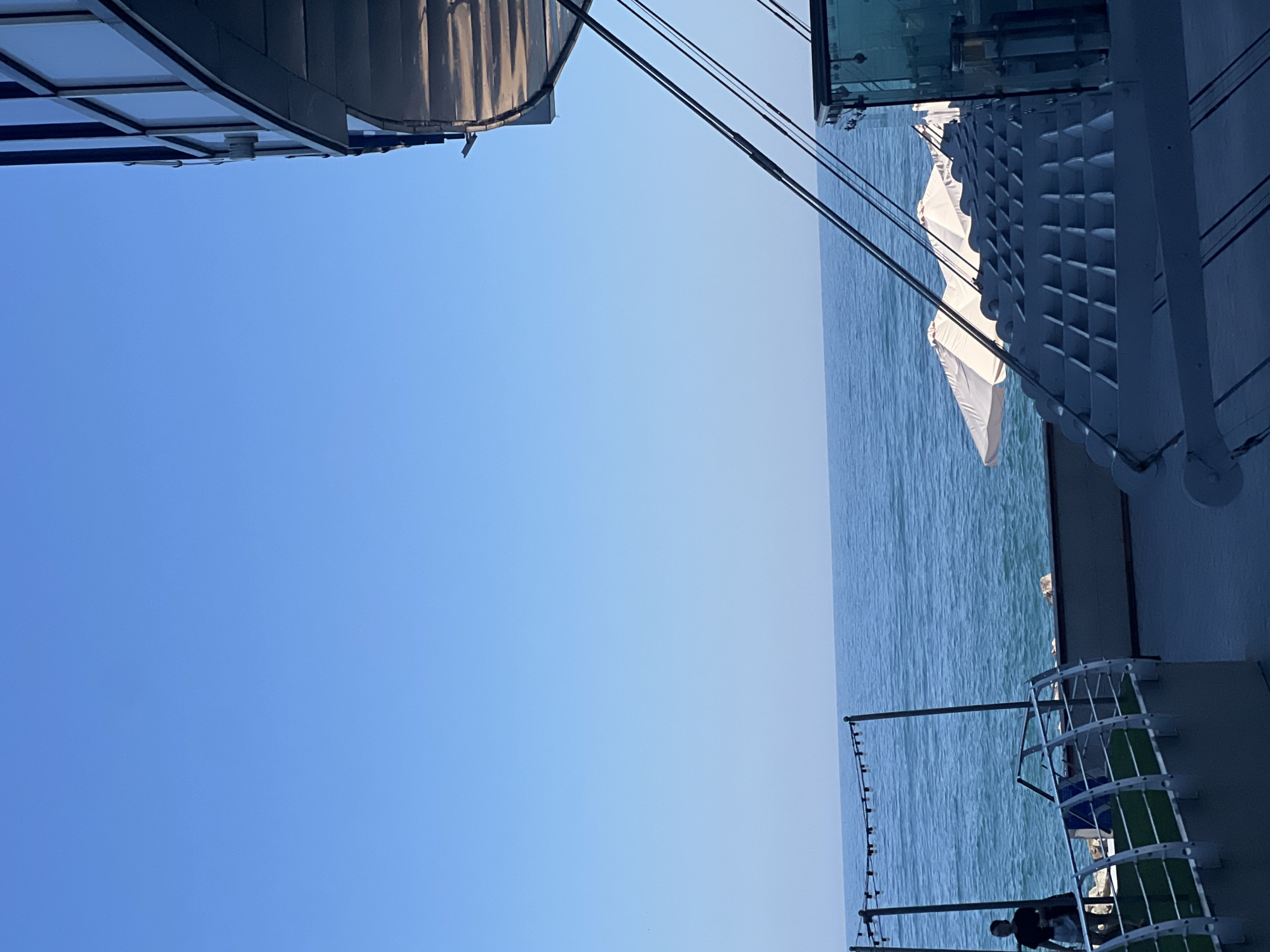 The image depicts a waterfront scene in Odesa, Ukraine. In the foreground, there is a modern structure with a curved balcony made of glass and metal. Below the balcony, a person is visible standing on a walkway next to the sea. Further out, white umbrellas are partially visible over a seating area, suggesting an outdoor cafe or restaurant. The sea is calm and blue, stretching to the horizon under a clear sky. In the distance, the coastline is lined with rocks. The overall impression is a serene and sunny day by the Black Sea.