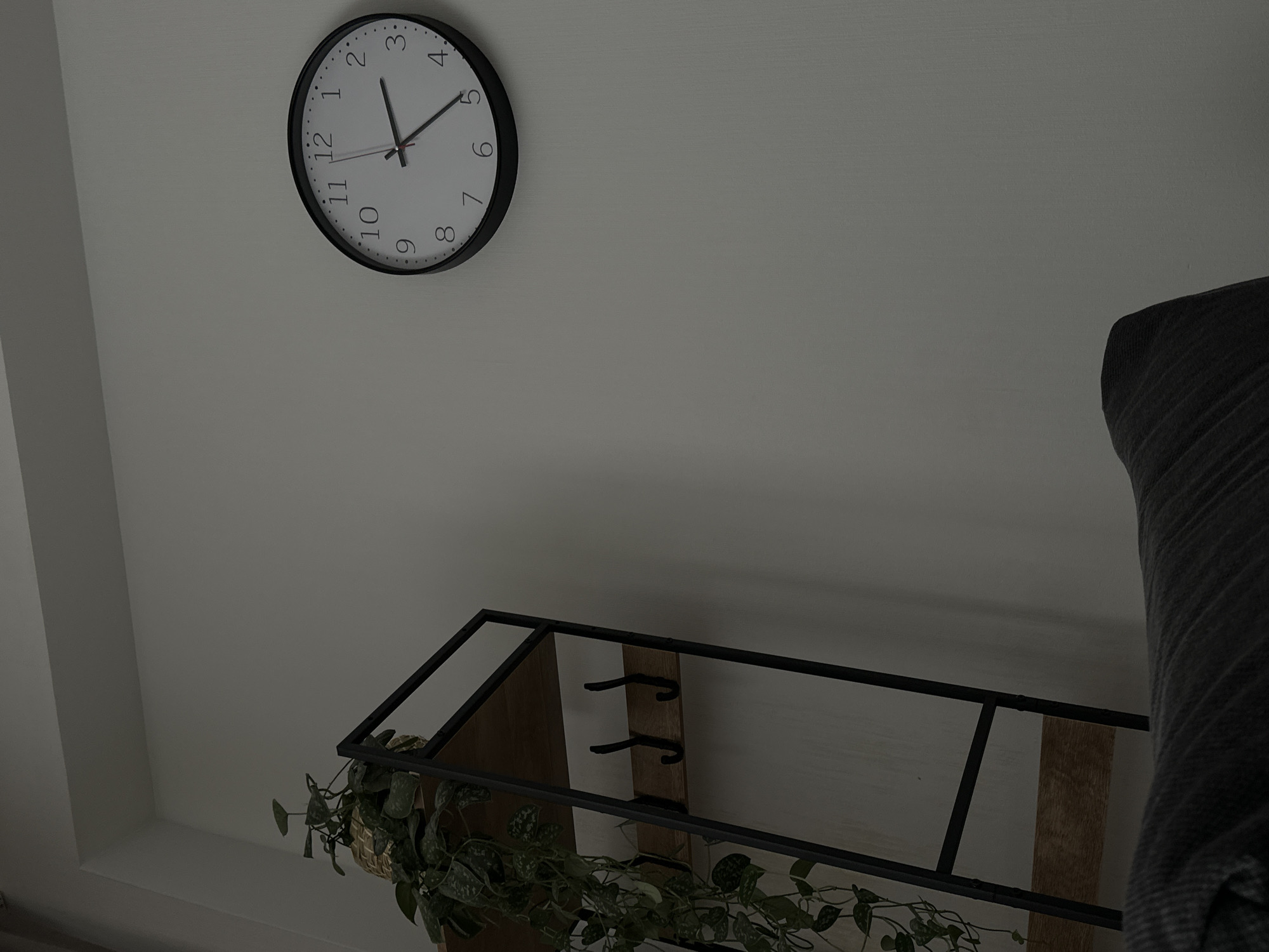 The image shows a high-angle view of an interior space. A round black clock with white numbers is mounted on a plain white wall, displaying a time of approximately 1:22. Below and to the right of the clock, a black metal shelf with wooden planks holds a potted plant with trailing vines. The shelf is positioned in the lower portion of the frame, and the corner of a gray upholstered furniture piece is visible to the right. The environment suggests a contemporary or minimalist interior setting. The location is not discernible from the image.