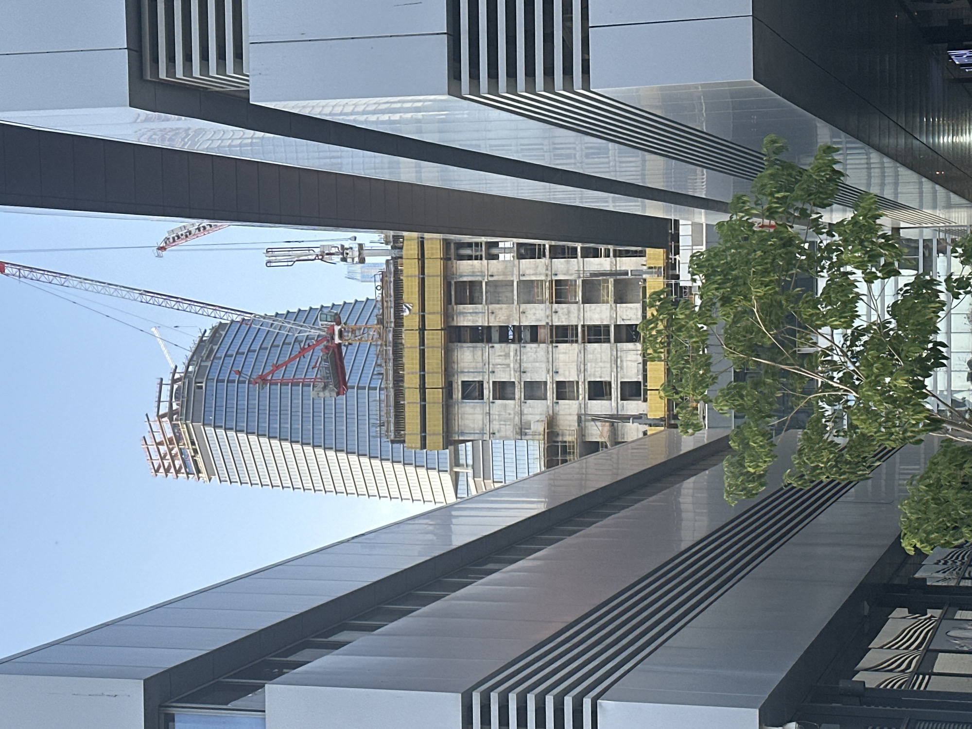 The image captures a view in Tel Aviv, Israel, showing a partially constructed high-rise building with a distinctive, curved facade. Construction cranes are visible near the building, indicating ongoing work. The foreground features architectural elements of another building, with a linear, geometric design in varying shades of gray. A tree with green foliage is also present. The sky is clear and blue.
