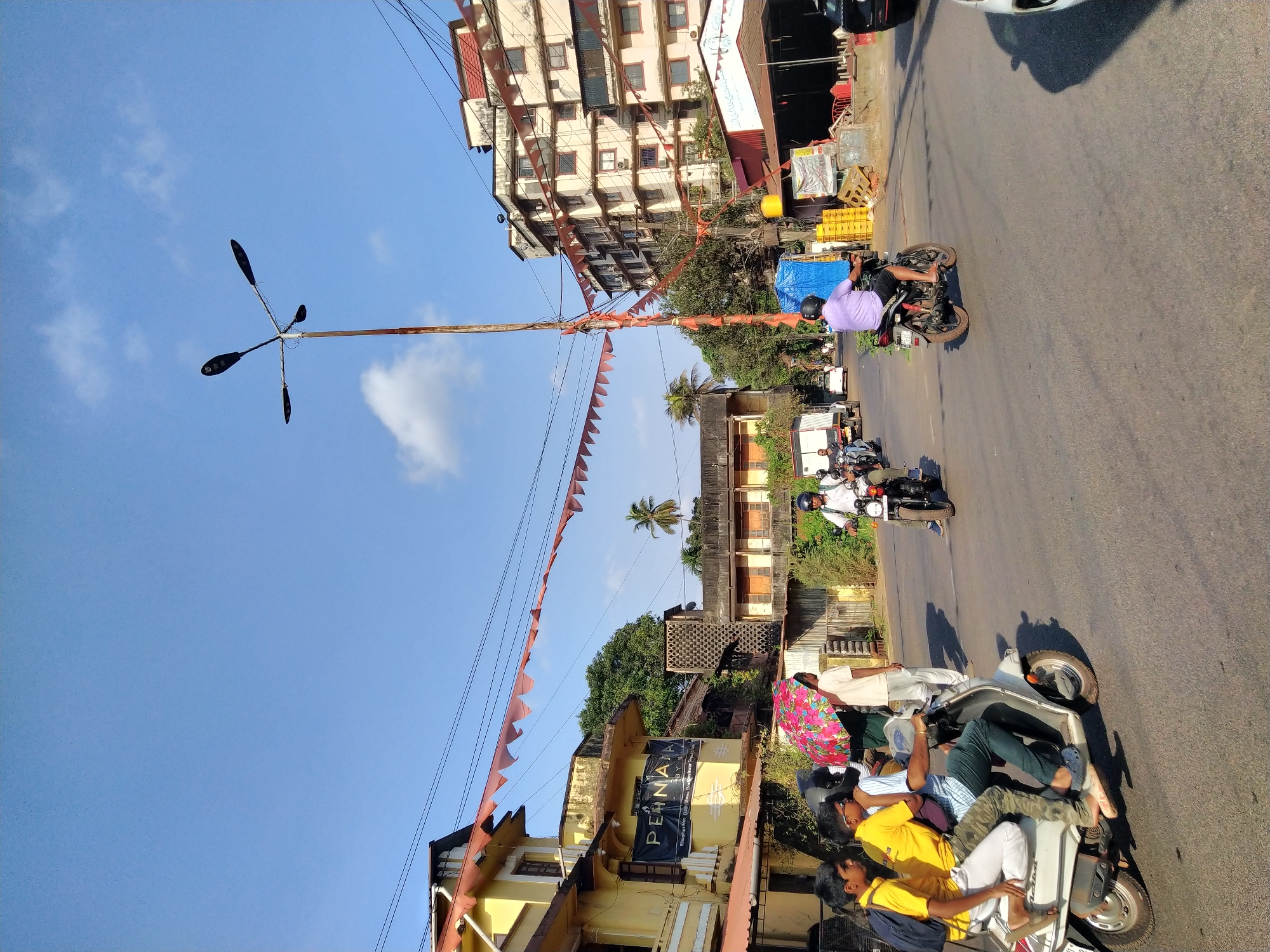 The image depicts a street in Mapusa, India, oriented vertically with the sky and a tall streetlamp on the left. The street is lined with buildings of varying heights and colors, including a taller, light-colored apartment building on the upper left and a yellow building with the name "PERMAJA" visible on its facade in the lower left. Red triangular pennants are strung across the street.
The street is occupied by several motorcycles and scooters. A scooter overloaded with several people is visible in the lower right corner, including one person holding a colorful umbrella. Other motorcycles with single riders are further up the street.
The street surface appears to be asphalt. The sky is blue with a few scattered clouds. Trees and other vegetation are interspersed among the buildings. Overhead wires are visible across the sky.