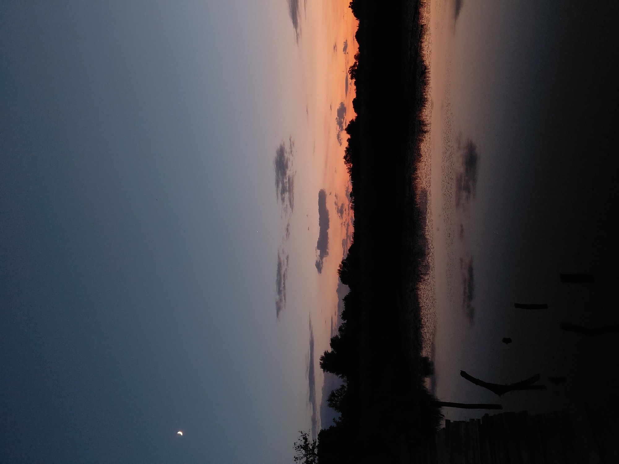 This image depicts a serene dusk scene on a body of water, likely a lake or river, in Ustyvytsia, Ukraine. The sky is a gradient of deep blue transitioning to a warm orange and pink horizon, indicating the sun has recently set. A sliver of a crescent moon is visible in the upper left quadrant of the sky. The silhouette of a dense tree line forms a dark border across the middle of the image. The still water perfectly reflects the sky and the dark treeline, creating a mirror image effect. In the foreground, on the left side, a portion of a wooden pier or walkway is visible, with a few dark, vertical posts emerging from the water. The overall atmosphere is peaceful and tranquil.