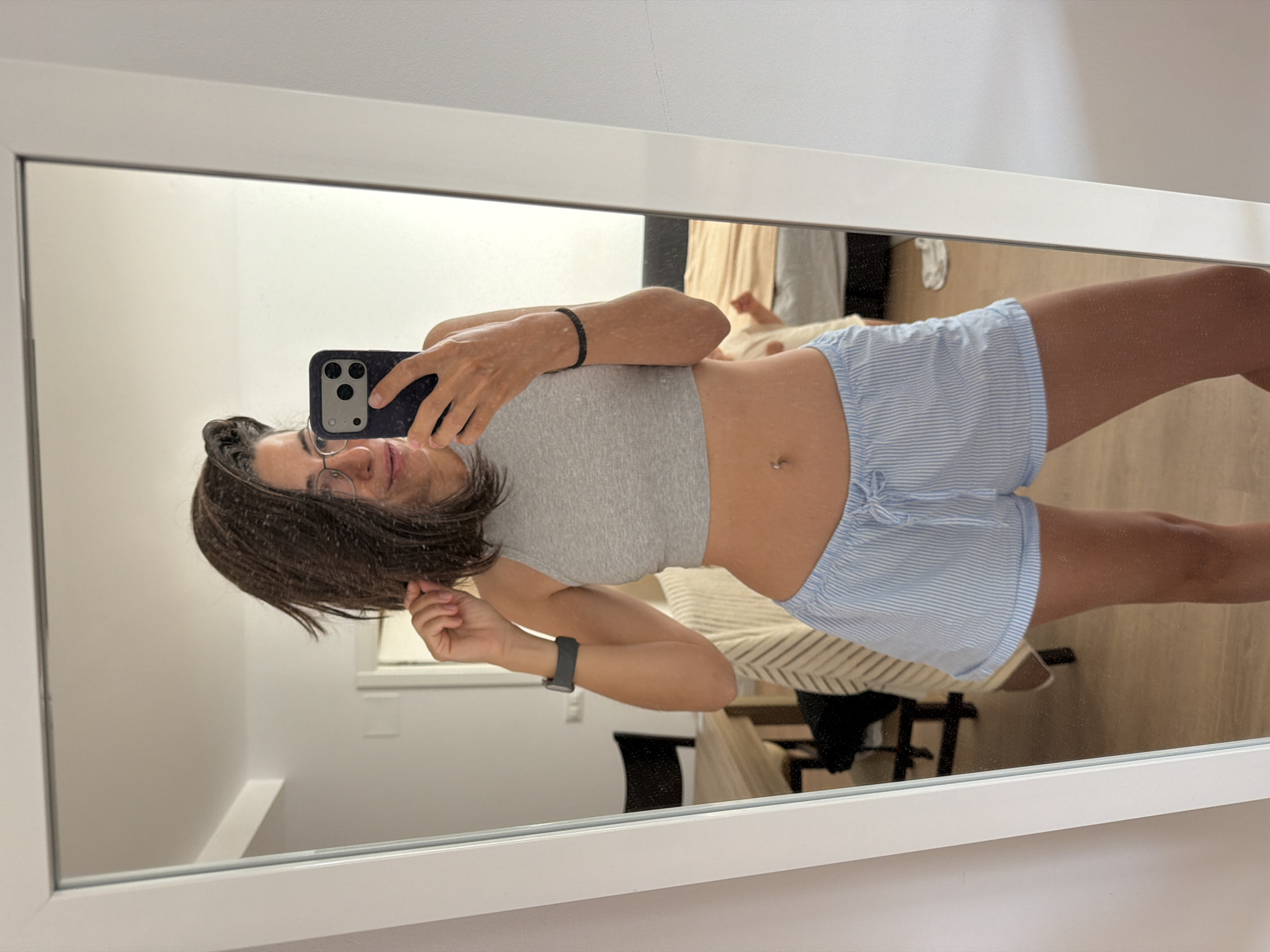 A person is taking a mirror selfie indoors. They are wearing a grey crop top and light blue and white striped pajama shorts. The person has dark hair and is wearing glasses. They are holding a smartphone in their right hand and adjusting their hair with their left hand. A smartwatch is visible on their left wrist. The background shows a room with a bed, a table with chairs, and a mirror. The image is taken in a residential setting. The location is l'Hospitalet de Llobregat, Spain.