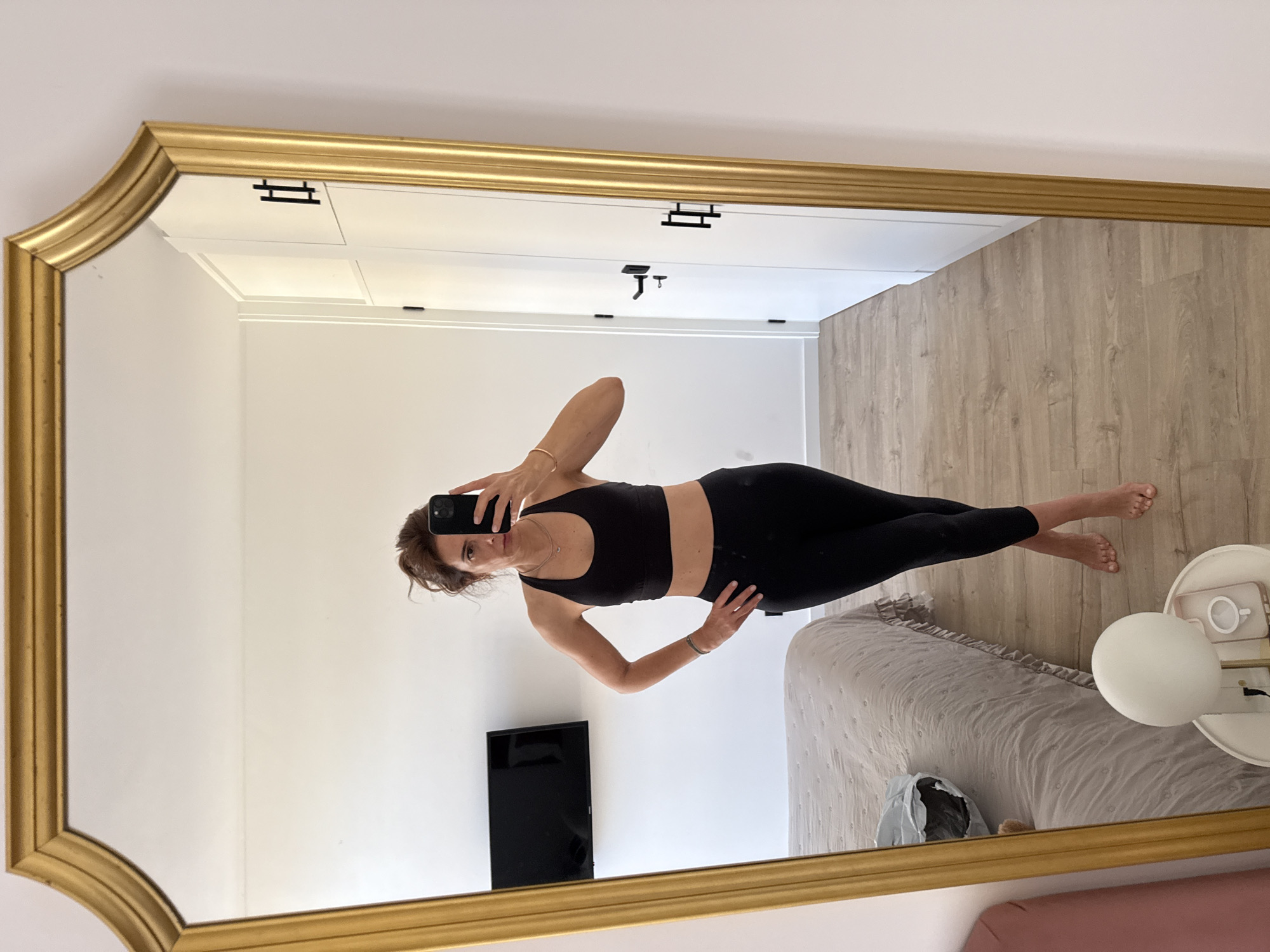 A woman in a black sports bra and leggings is taking a mirror selfie in what appears to be a bedroom. The mirror has a gold frame. Behind her in the mirror, a bed is visible. There's also a small television mounted on the wall. The room is well-lit and has light-colored walls and flooring. Based on the interior architecture and style, it is not possible to determine its location in Vila Nova de Gaia, Portugal from this image. The objects on the bedside table are likely a lamp, a phone, and a coffee mug. The woman is holding a smartphone to take the photo, and her arm is slightly bent.