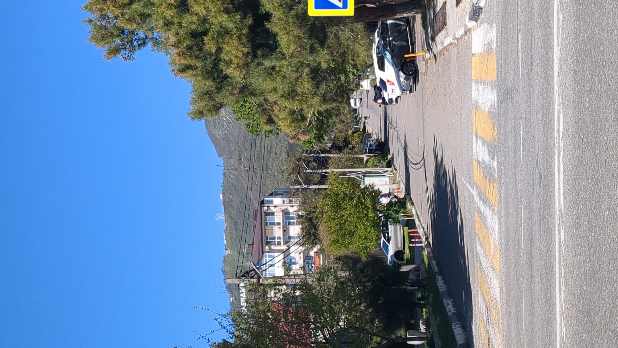 This media file presents a bright, sunny street scene in Gelendzhik, Russia. The image is taken from the perspective of a person standing on or near the street, looking down a road that curves slightly to the left.
The main subjects include an asphalt road with clear yellow and white zebra crossing markings, indicating a pedestrian crosswalk. Several cars are parked along the right side of the road, with a prominent white car in the foreground. Abundant green trees and bushes line both sides of the street, with some trees casting long shadows across the road, suggesting it is either early morning or late afternoon. Utility poles and overhead wires are visible, running parallel to the street.
In the mid-ground, multi-story buildings with balconies are nestled among the foliage. In the far background, a large, tree-covered mountain range dominates the horizon under a clear blue sky. A faint circular structure, possibly a Ferris wheel, can be discerned on the mountain slope.
A notable detail is the visible text on a building sign: "АРМЯНСКИХ КОНЬЯКОВ" (Armenian Cognacs), with the word "ШАХ" (SHAKH) also decipherable, indicating a business selling Armenian cognacs. The weather is clear and sunny, and no people are visible, giving the scene a quiet and calm ambiance.