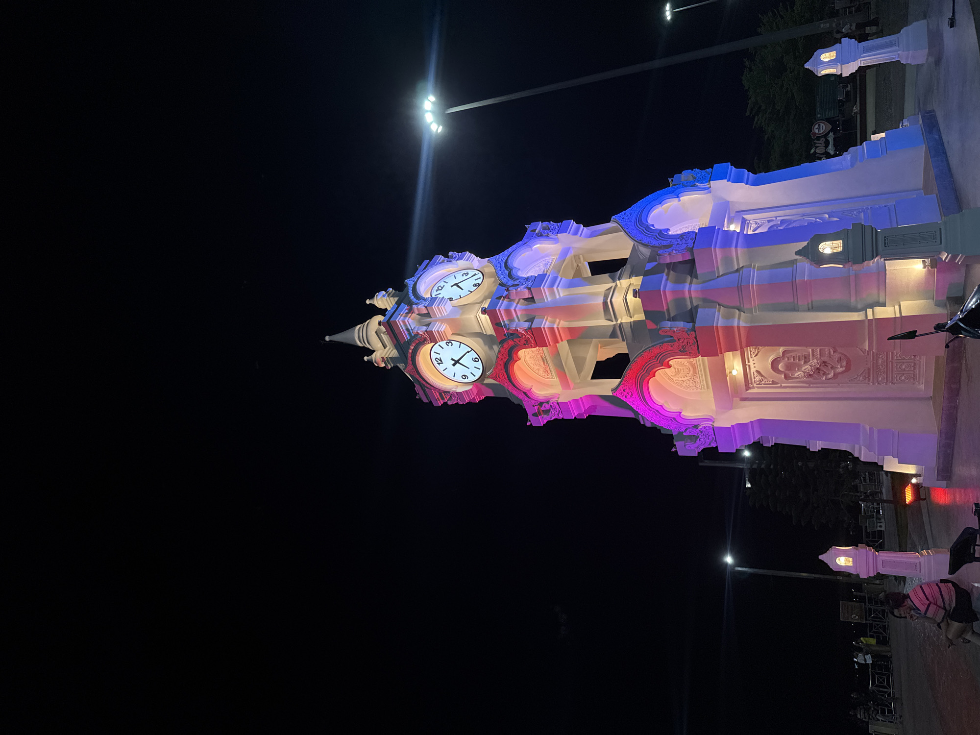 An ornate, multi-tiered clock tower, illuminated with vibrant pink and blue lights, stands prominently in the foreground of a night scene. The tower features decorative carvings and multiple clock faces showing the time. To the left of the tower, a few individuals are seated and standing. In the background, streetlights and trees are visible against a dark sky. The location is the Surat Thani City Municipality Clock Tower in Surat Thani City, Thailand.