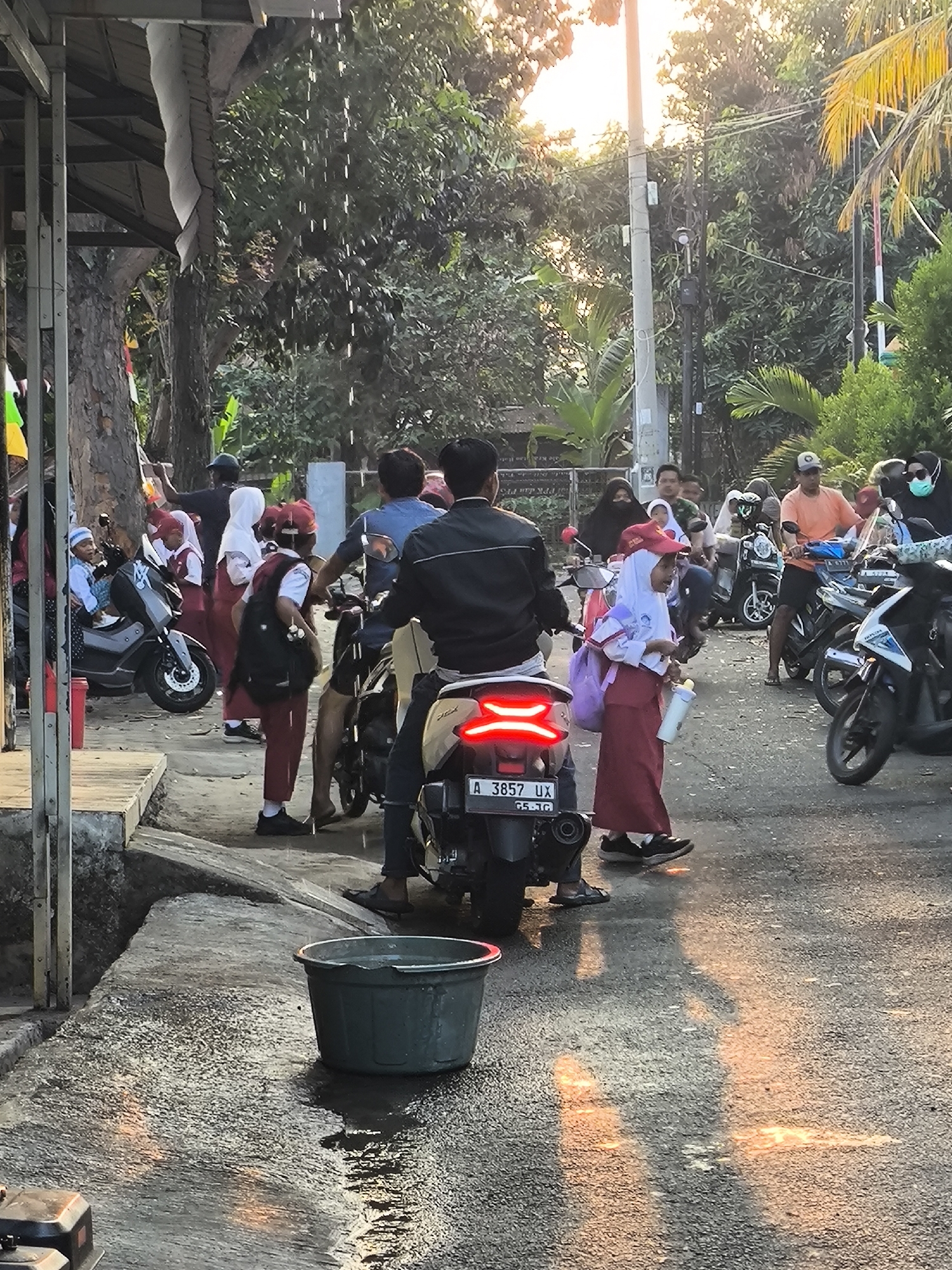 A group of people, some in school uniforms, are gathered around motorcycles on a street in Cilegon, Indonesia, likely during the late afternoon due to the warm, golden light filtering through the trees. The scene appears to be a common occurrence in a residential or public area, possibly near a school or market. Water is dripping from an overhang on the left side of the image, creating a wet patch on the pavement. A large green tub filled with water is also present on the ground. The people are dressed in a mix of casual wear and school uniforms, with some wearing hijabs. Several motorcycles are parked or being ridden, indicating transportation is a key element of the scene. The overall atmosphere is one of everyday life in a bustling Indonesian city.