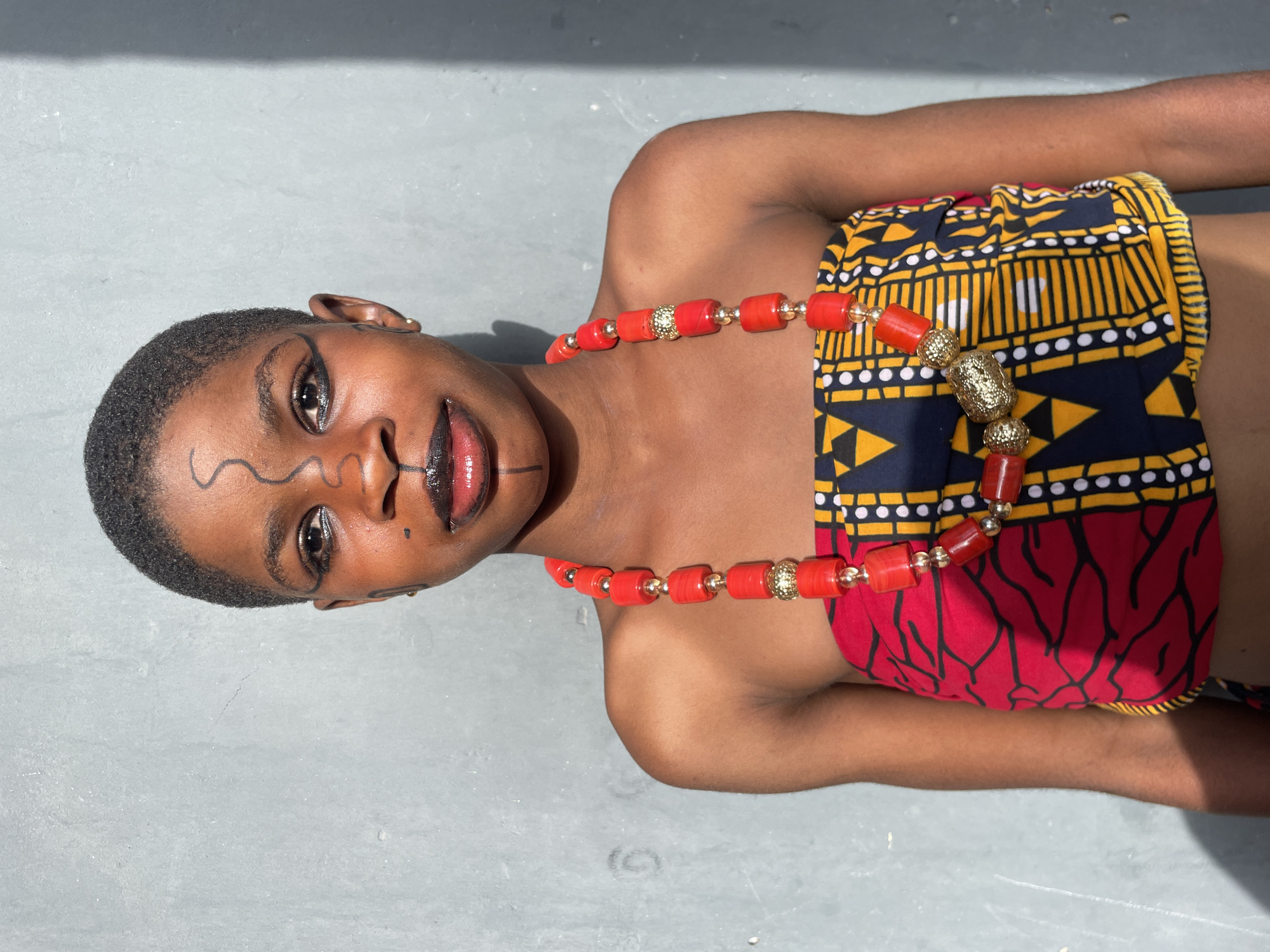 A captivating portrait features a young, dark-skinned individual with closely cropped hair, lying on a light gray, textured surface, likely concrete, under bright sunlight. The person is adorned in vibrant traditional African attire and jewelry, reflecting the cultural context of Badagry, Nigeria.
They are wearing a strapless top or wrap garment made from Ankara fabric, divided into two distinct patterns. The upper portion, covering the chest, showcases a dark blue background with striking yellow and black geometric designs, including stripes, triangles, and white dots. The lower section, around the midriff, transitions to a rich red background adorned with intricate black, organic, tree-like or abstract patterns.
A prominent necklace graces their neck, composed of chunky, cylindrical red beads interspersed with ornate, textured golden spherical beads. The individual's face is intricately decorated with black lines: a wavy design across the forehead and another below the left eye, along with a line extending from the right side of the mouth. Their lips are painted with a dark, glossy shade, possibly black or deep red, and there's a small dot below the right eye. A small, gold-colored earring is visible on their left ear.
The subject's eyes are open, meeting the viewer's gaze with a calm and direct expression. The bright, natural sunlight highlights their features and the glossy elements of their makeup, while casting subtle shadows that add depth to the portrait. The scene is a thoughtfully posed, outdoor shot, emphasizing cultural identity and artistry.