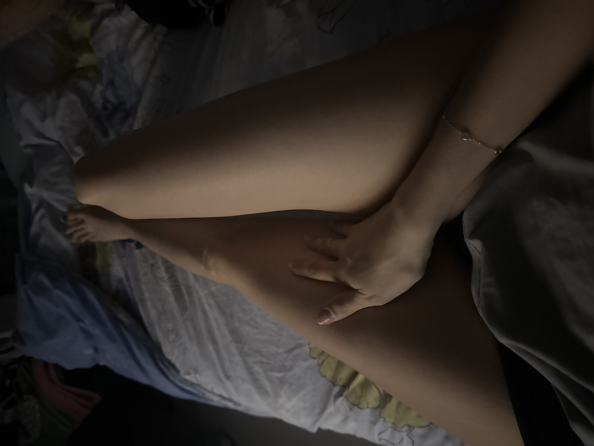 A close-up, dimly lit, high-angle shot shows a person lying on a bed. The person's legs are visible, with one leg crossed over the other. A hand rests on the top leg. A delicate bracelet adorns the wrist of the arm crossed over the legs. The bed is covered with wrinkled sheets and blankets. The location and city of Izhevsk, Russia are not discernible from the visual information provided.