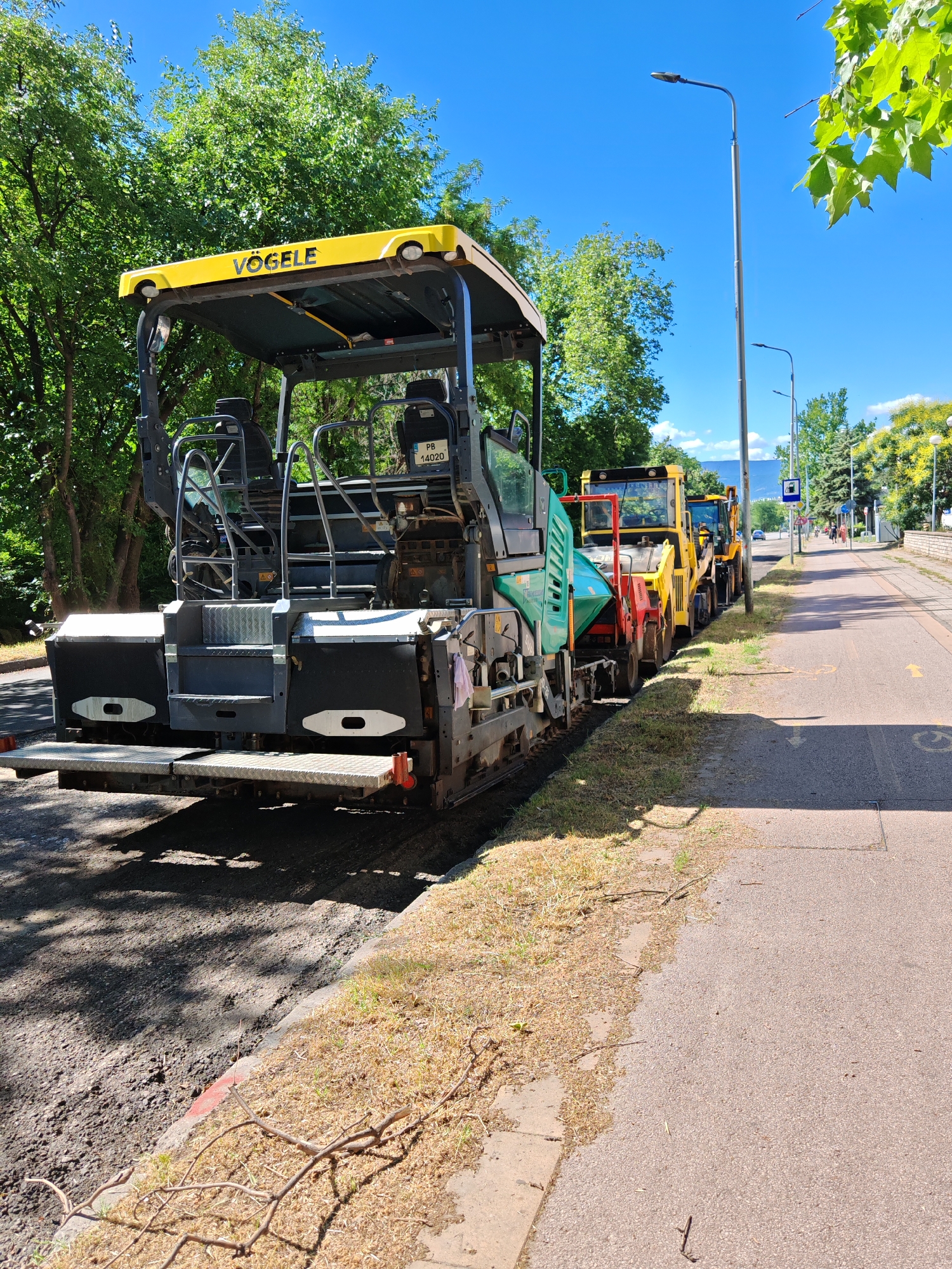 The image depicts a road construction scene on a sunny day, likely in Plovdiv, Bulgaria, given the presence of construction equipment commonly used for such projects in European cities. In the foreground, a large yellow and gray VOGELE asphalt paver is positioned on a freshly laid asphalt surface. Behind it, a line of yellow construction vehicles, including excavators and a loader, are parked alongside the road. The area is lined with trees and a pedestrian walkway with yellow markings is visible to the right. The sky is clear blue.