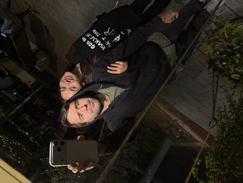 The image depicts a man and a woman in an outdoor urban setting, identified as Tel-Aviv, Israel, at night. The man, positioned in the foreground and appearing to take a selfie using a light-colored smartphone held in his right hand, has short dark hair, light skin, and a facial expression characterized by wide eyes and a slightly open mouth. He wears a dark, zipped-up hooded sweatshirt over a green t-shirt and dark pants, with light-colored sneakers visible. A red bracelet is on his left wrist. His left arm is around the woman's shoulder.
The woman stands to the man's right, smiling towards the camera with her head tilted slightly. She has long dark hair and light skin. She wears a black hooded sweatshirt with white, mirrored text that appears to read "IN GOOD WE TRUST NO CHAOS WE WANT PEACE" and dark pants. Her right hand is placed on the man's left arm, displaying a ring on her ring finger.
The background is dark and blurred, illuminated by string lights overhead, suggesting an outdoor restaurant, cafe, or public plaza. Several indistinct figures are visible further back, some seated at what appear to be tables, and a wooden bench is also partially visible. The immediate foreground includes dark foliage, a low brick wall with some red markings, and a metal chain, all contributing to the outdoor urban environment.