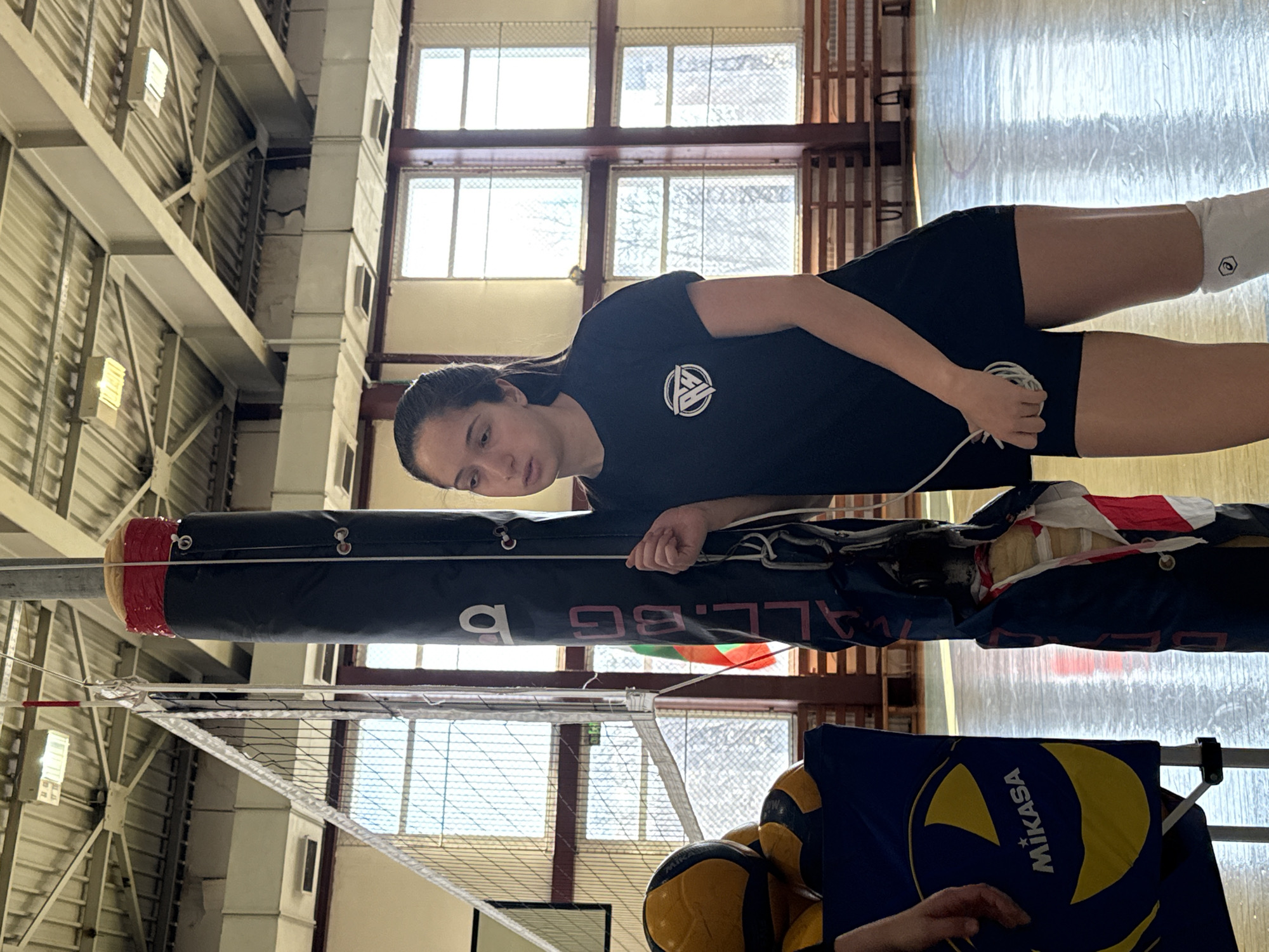 The image depicts a young woman standing in a gymnasium next to a volleyball net. The net is supported by a black pole with the word "BALL.BG" printed on it. Adjacent to the pole are several volleyballs and a blue Mikasa volleyball bag. The woman is wearing a dark-colored t-shirt with a logo on the chest, dark shorts, and a white compression sleeve on her right leg. She appears to be adjusting or checking the net's tension with her hands. The gymnasium features a high ceiling with exposed metal support structures and large windows that provide natural light. Based on the writing on the net's pole, the location is likely in Sofia, Bulgaria.