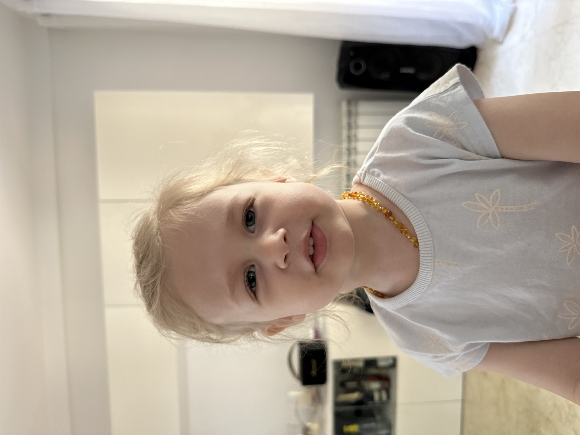 A young blonde child with blue eyes is standing in front of a wall. The child is wearing a light blue t-shirt with a palm tree pattern and an amber necklace. In the background, there are white cabinets, a radiator, and a window with white curtains. To the left of the child, a shelf unit holds several bottles and a small black box. To the right of the child, a black speaker is visible. The scene appears to be indoors, likely in a residential setting, given the presence of furniture and household items. The overall lighting suggests it is daytime. The location of Sant Feliu de Guíxols, Spain is not visually discernible from the provided image.